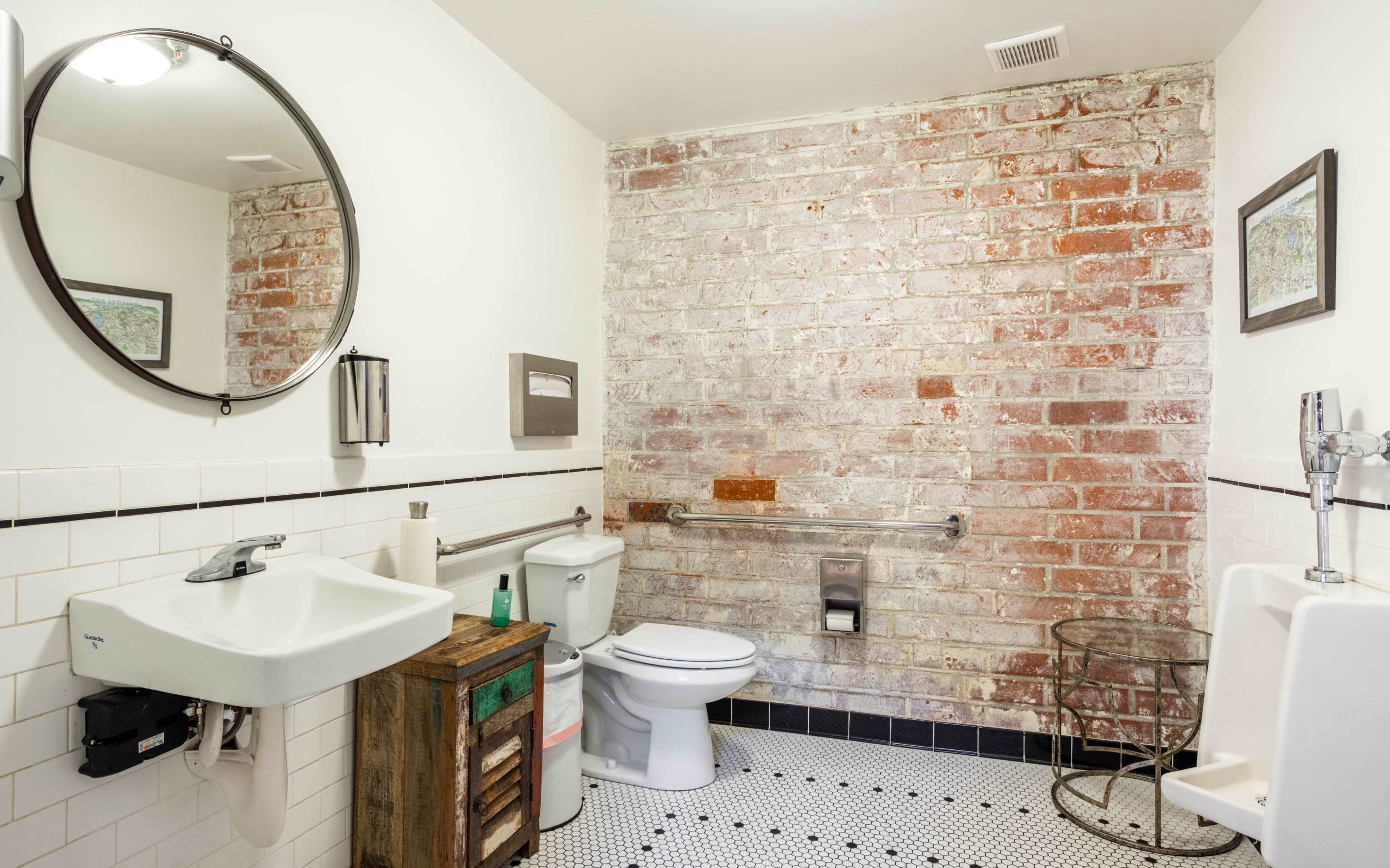 A rustic-style bathroom featuring exposed brick walls, a circular mirror, a white sink, a toilet, and a tiled floor with black and white patterns.