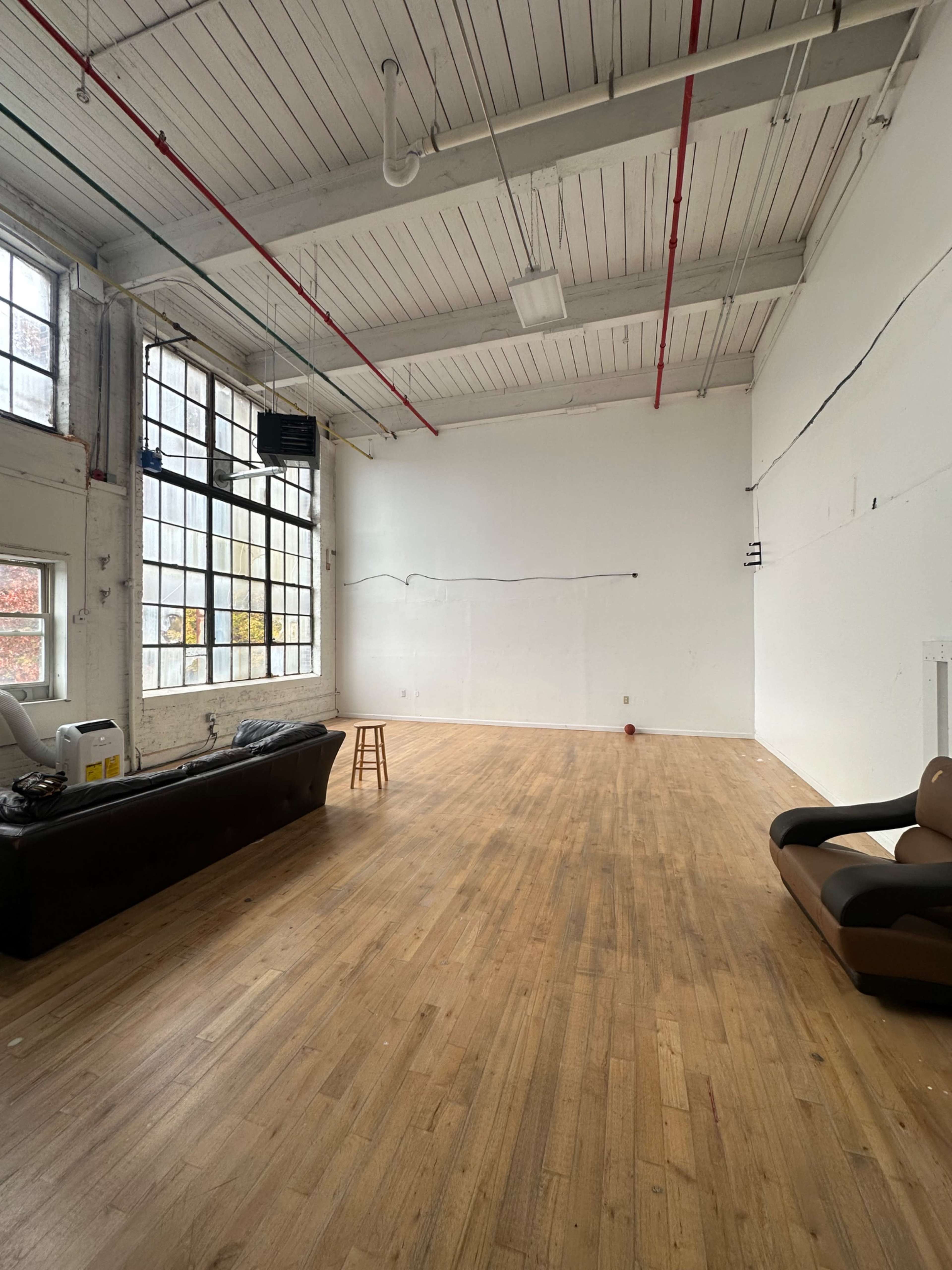 The image shows a spacious, empty room with wooden flooring, large windows, and minimal furnishings, including a couch and a stool.