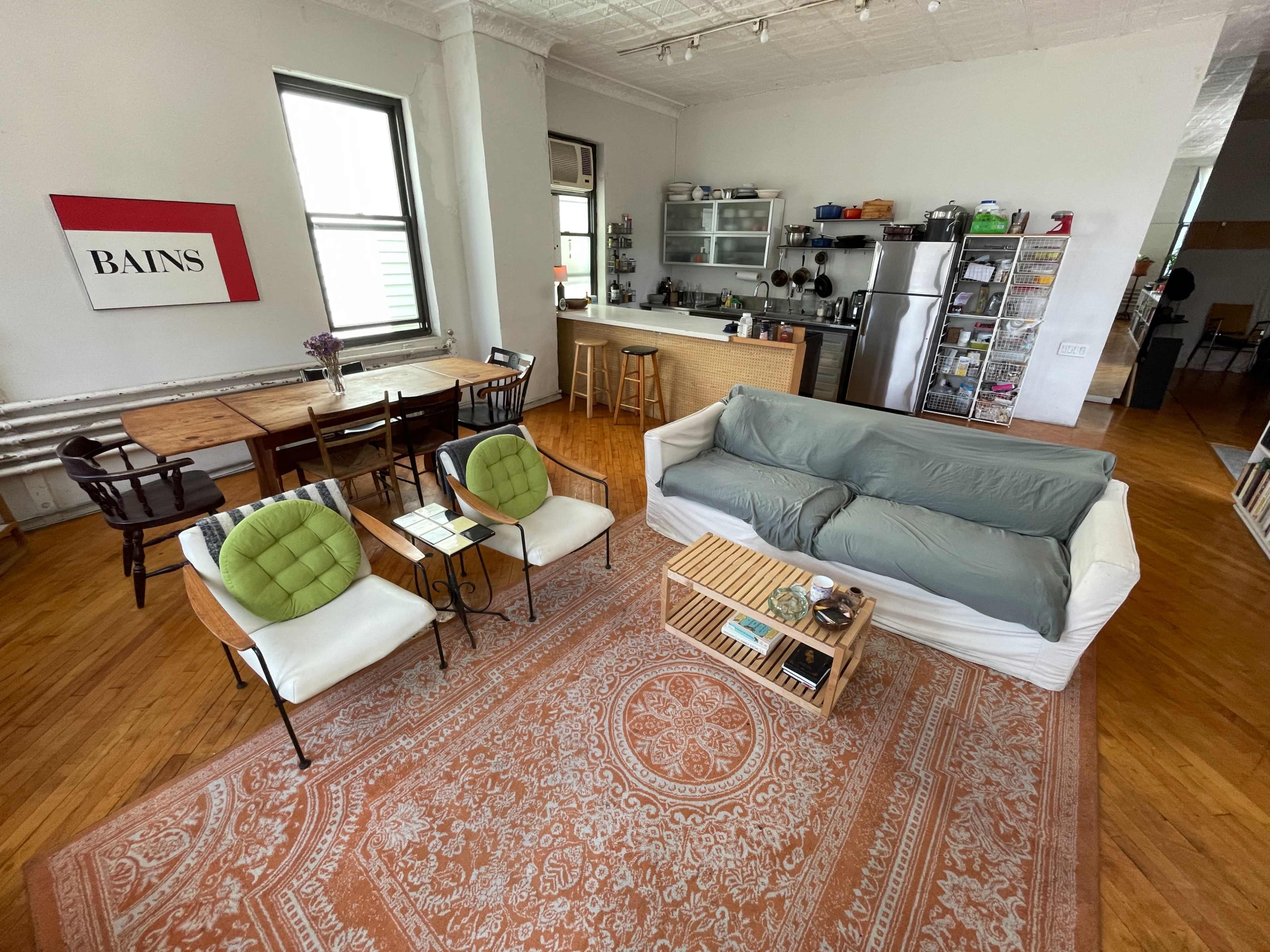 The image shows a spacious, modern living area with a large rug, a sectional sofa, two chairs, a wooden dining table, and an open kitchen in the background.
