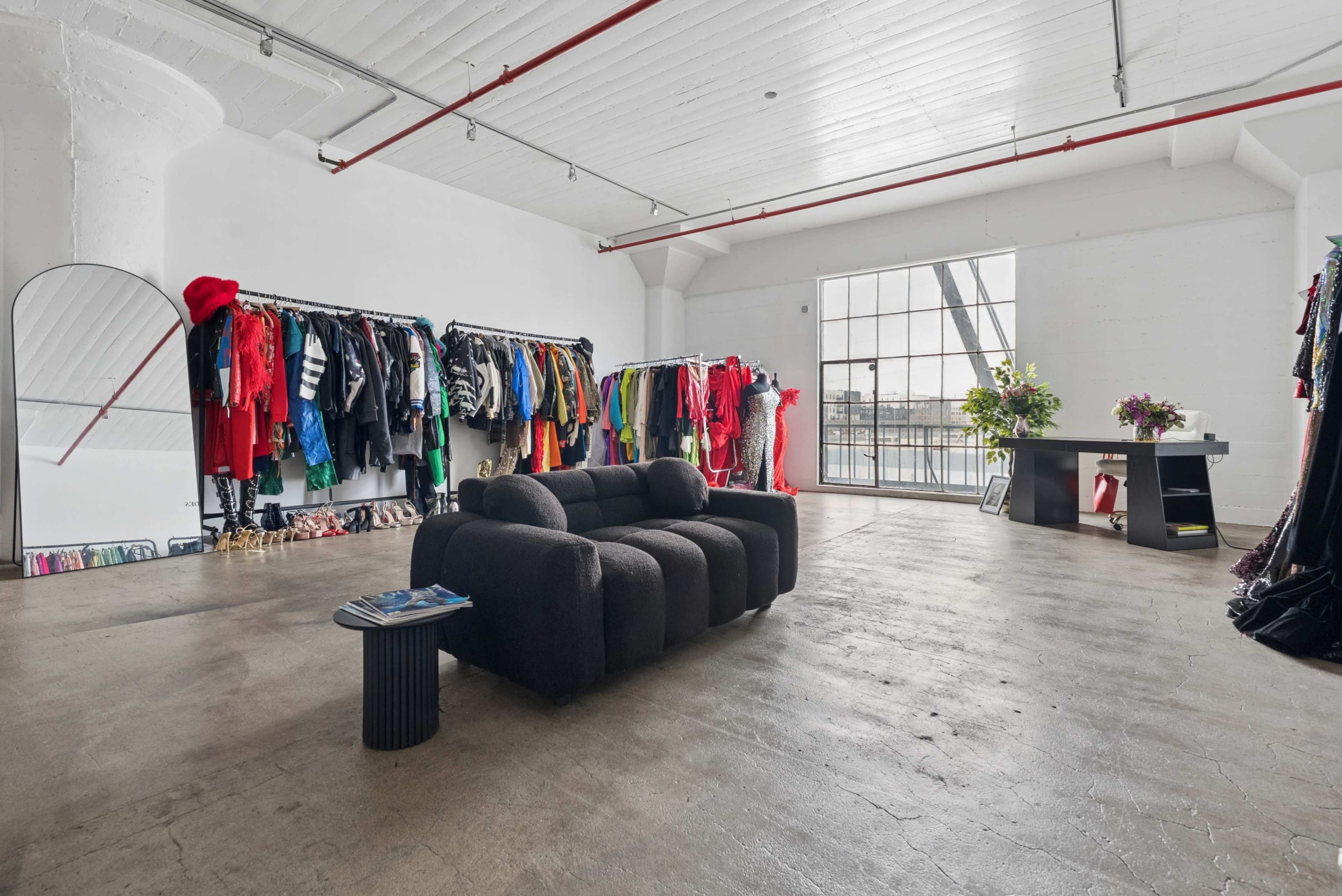 The image shows a spacious interior with a black couch in the foreground, clothing racks filled with colorful garments along one wall, and large windows letting in natural light.