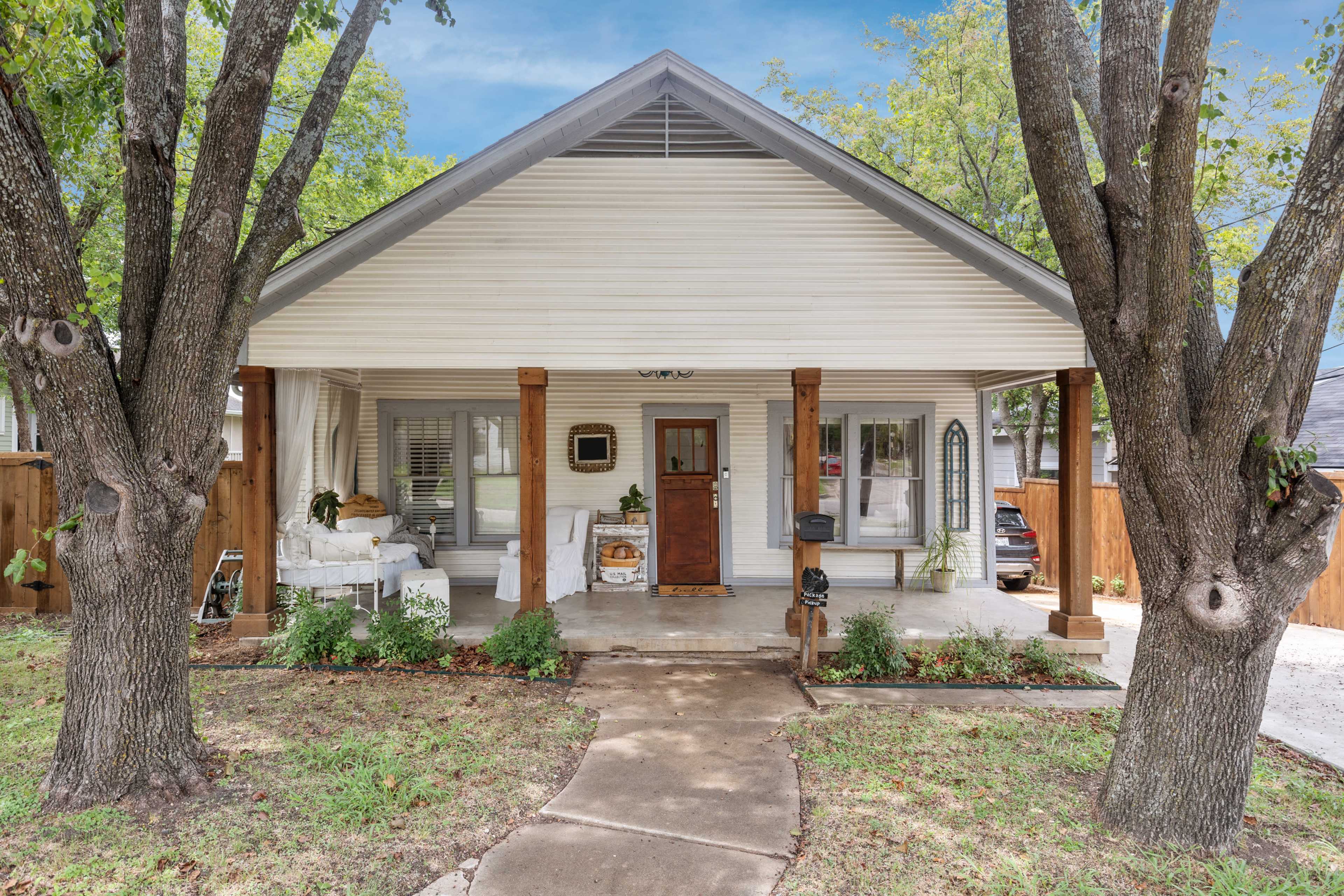 A single-story, white paneled house with a covered porch, flanked by two large trees and a pathway leading to the front door.