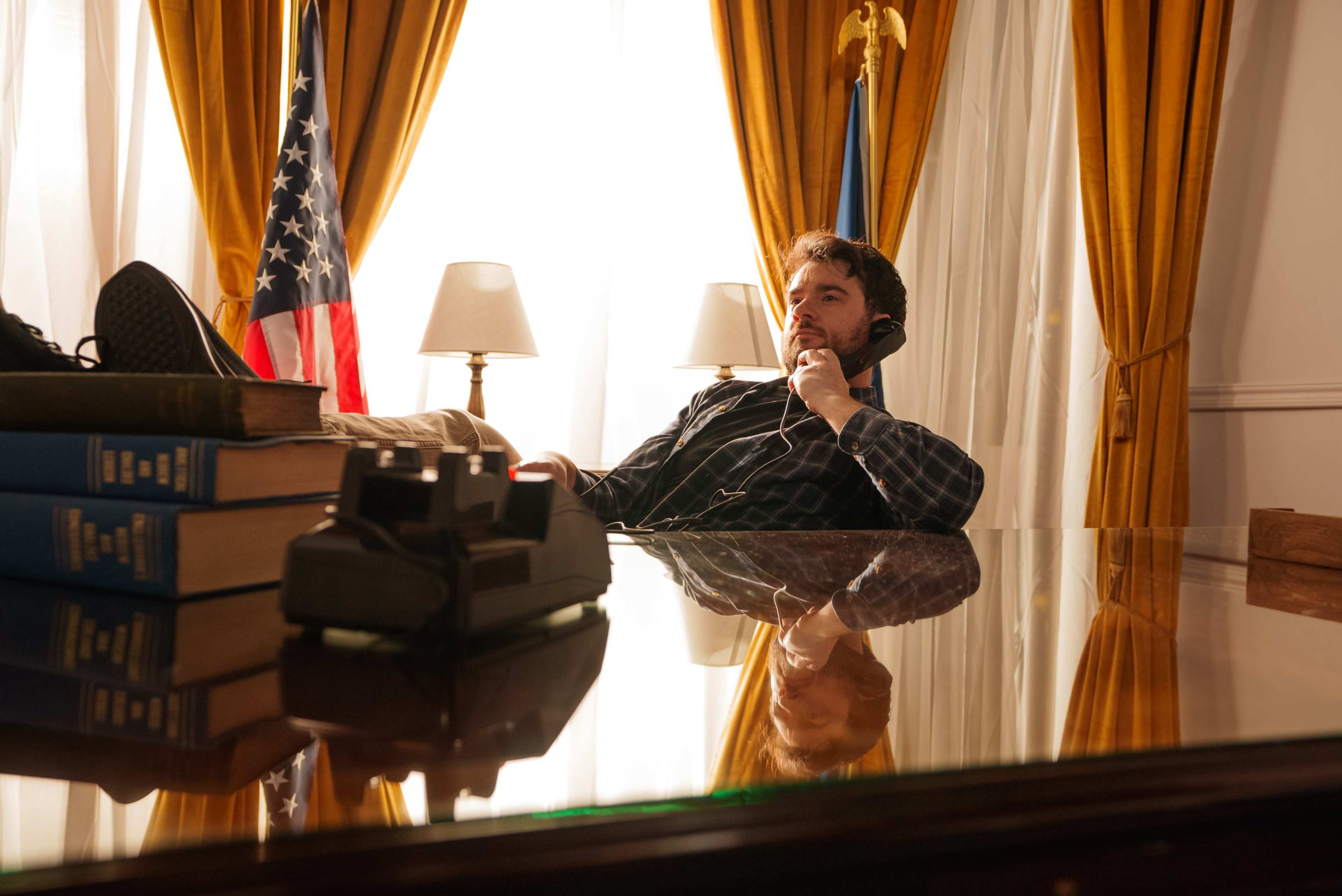 A man in a checkered shirt rests his feet on a desk while wearing headphones in a room with an American flag and yellow curtains.