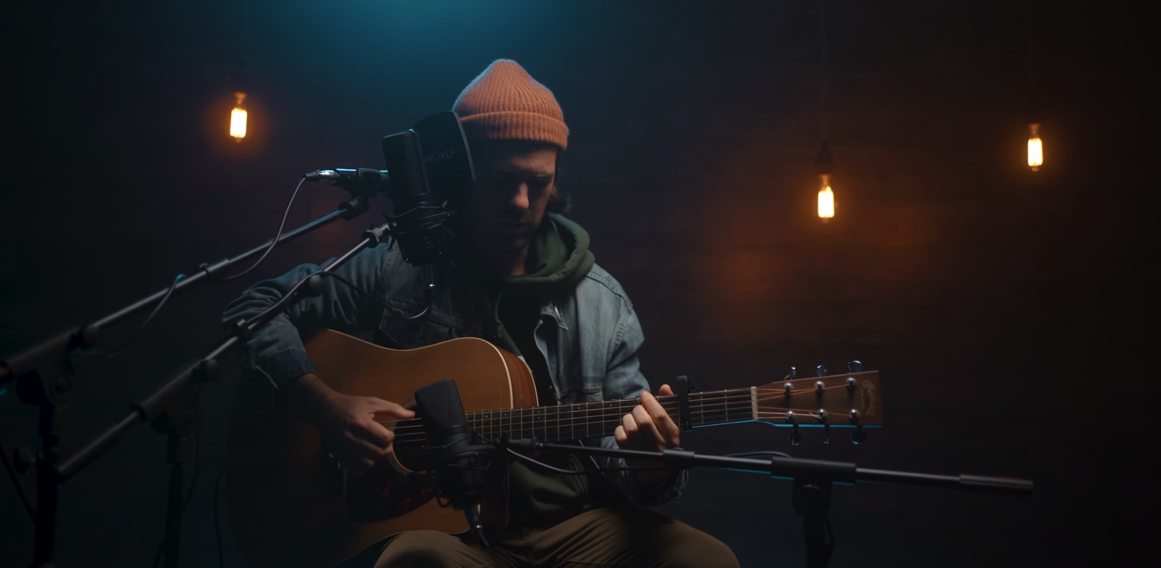 A musician sits on stage with an acoustic guitar, wearing a beanie and a denim jacket, surrounded by dimly lit hanging lights.