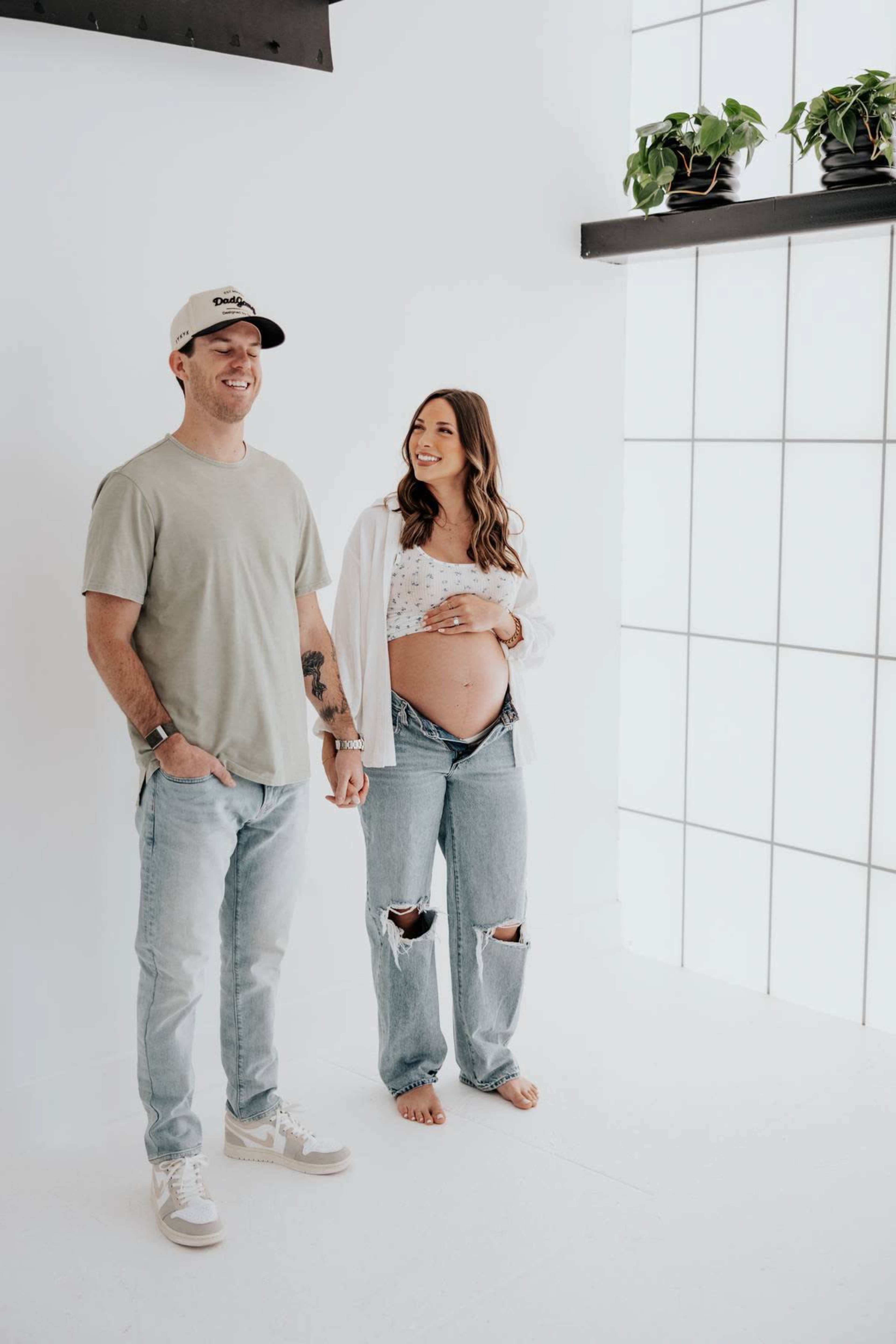 A couple stands together in a bright studio, with one partner holding their pregnant belly while smiling at the camera.