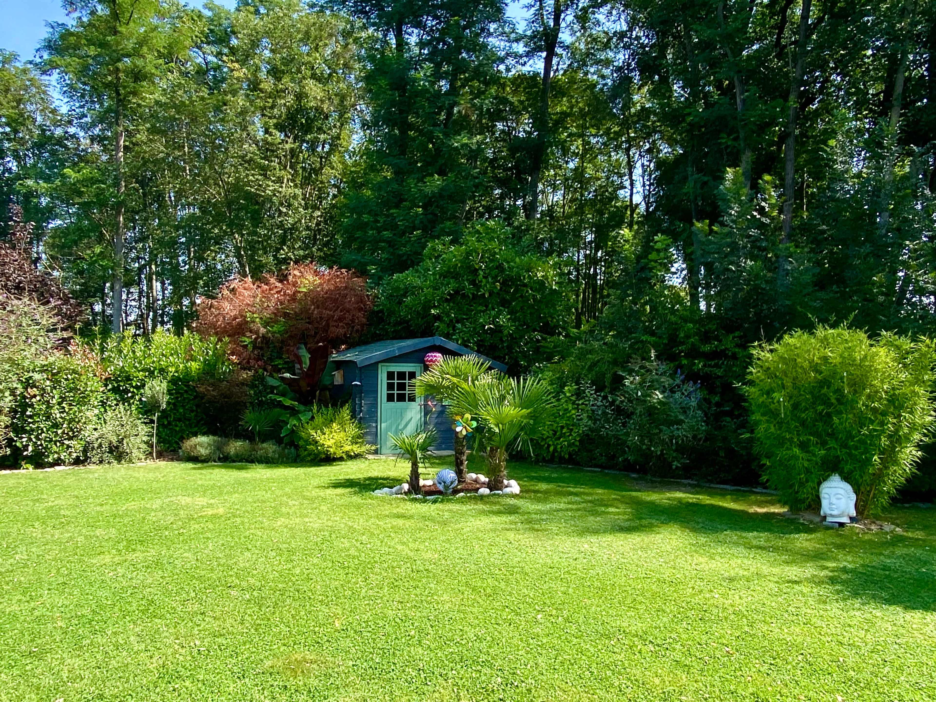 A well-maintained garden features a small green shed surrounded by various plants, bushes, and a stone statue in a serene setting.