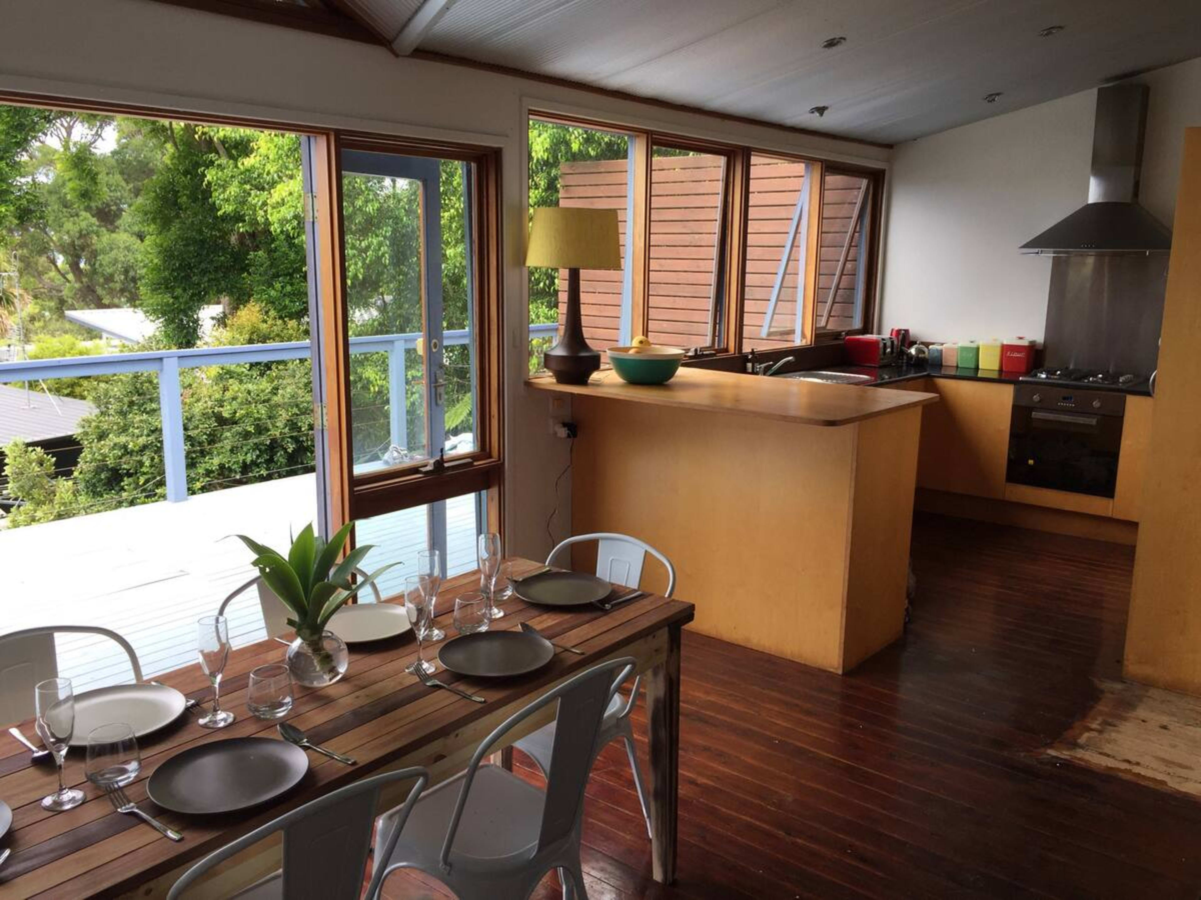 A modern kitchen with wooden flooring features a dining table set for six and large windows allowing natural light to fill the space.