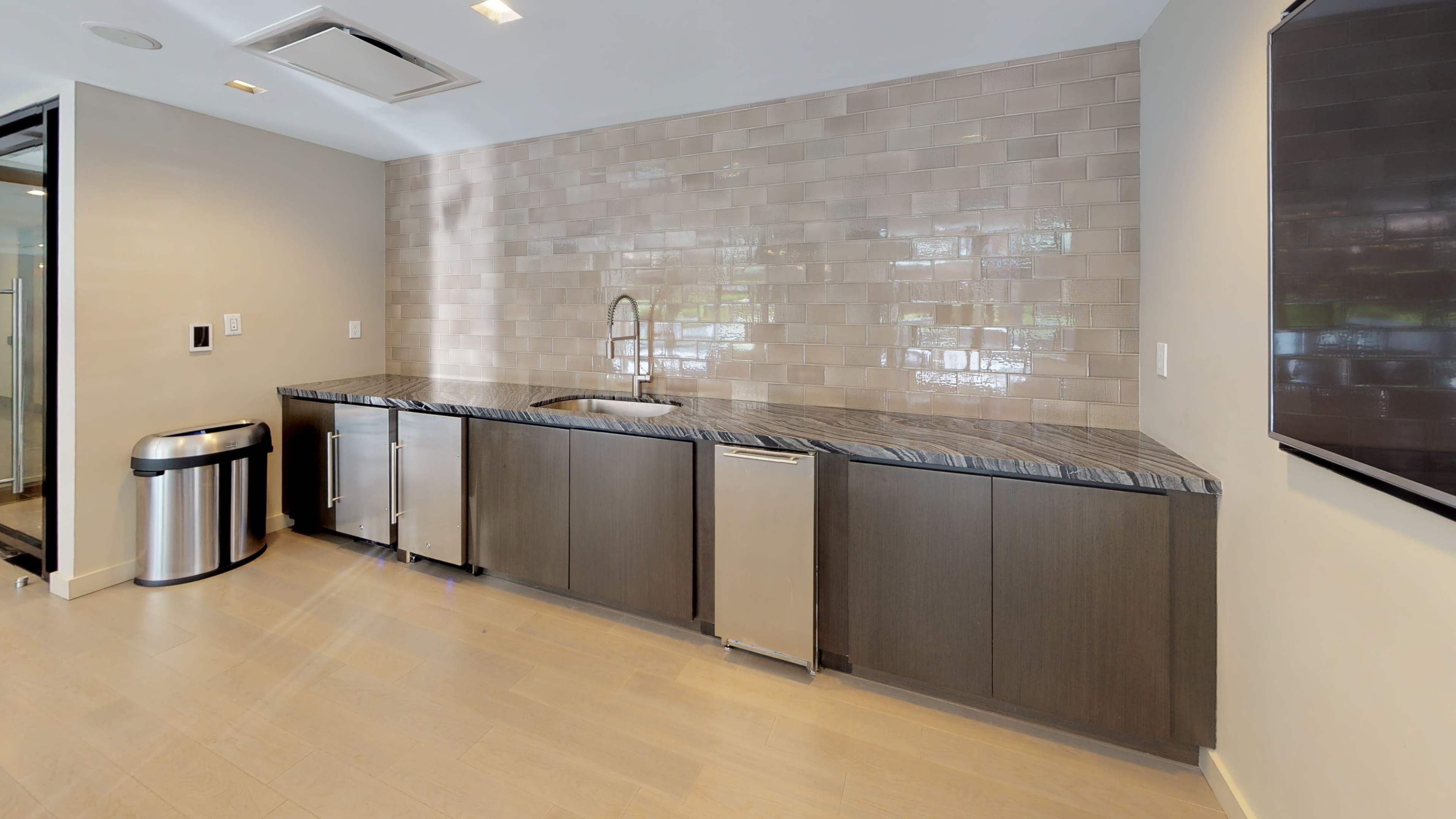 A modern kitchen features a granite countertop with a sink, dark cabinets, and a stainless steel refrigerator, along with a trash can and wall-mounted television.