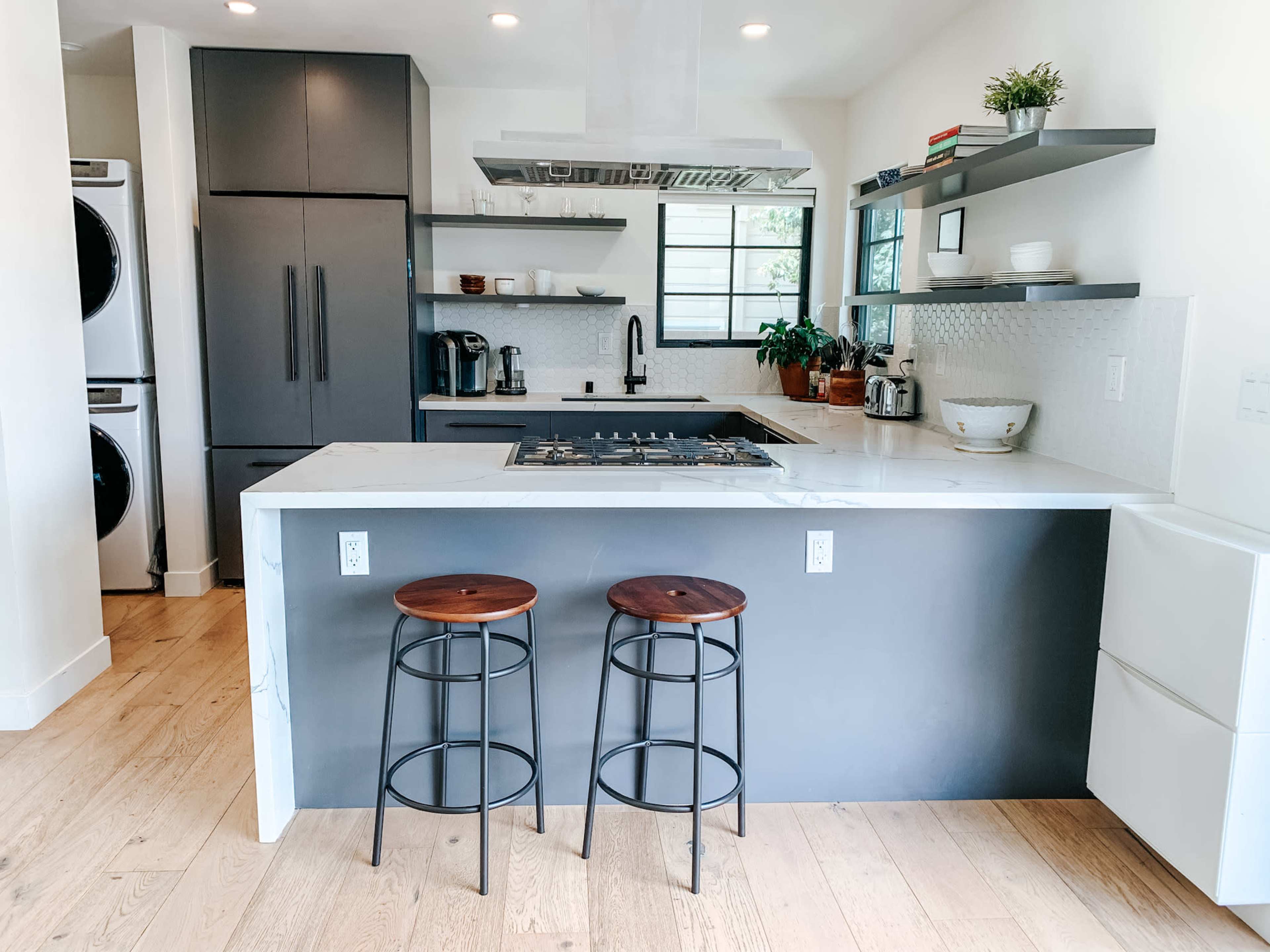 A modern kitchen features a central island with two wooden stools, sleek black cabinetry, and stainless steel appliances.
