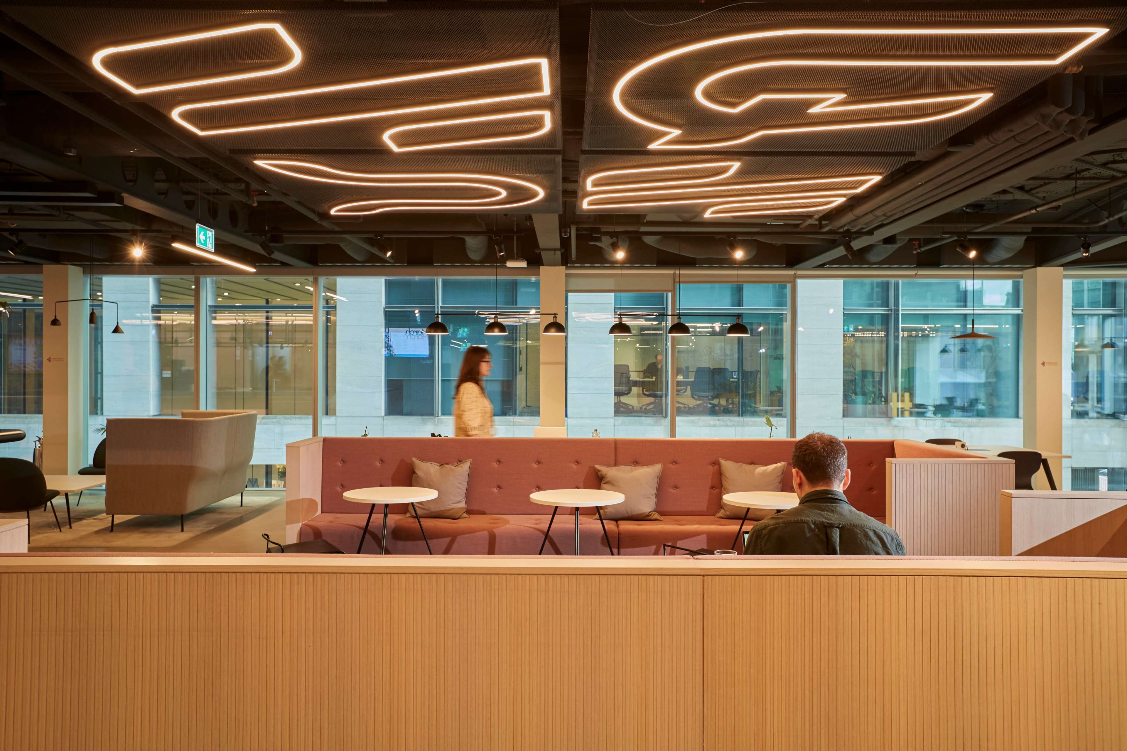 A modern office space features a low pink seating area with round tables, surrounded by large windows and illuminated by abstract ceiling lighting.