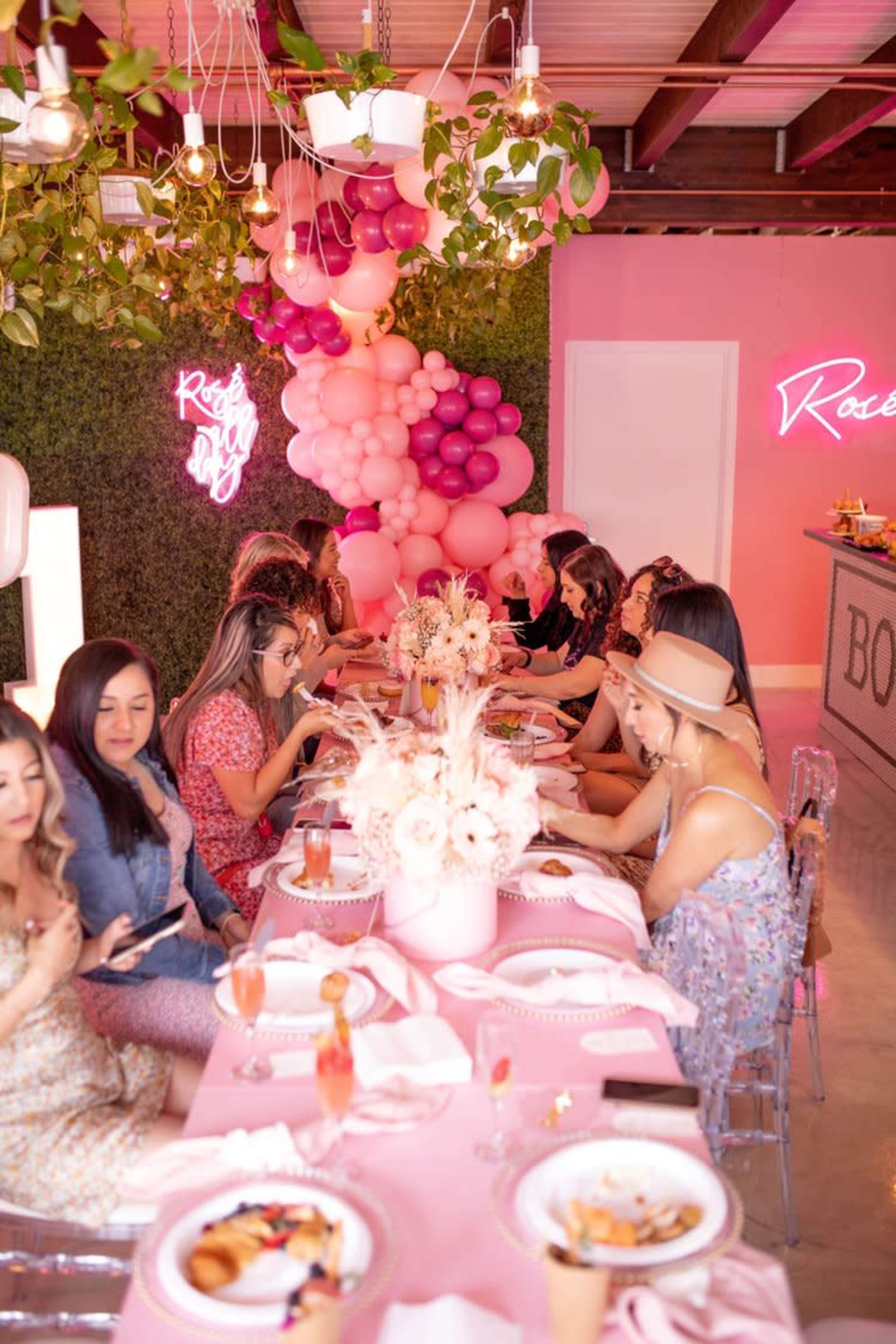 A group of women is seated at a long table adorned with pink decorations and floral arrangements in a colorful event space.