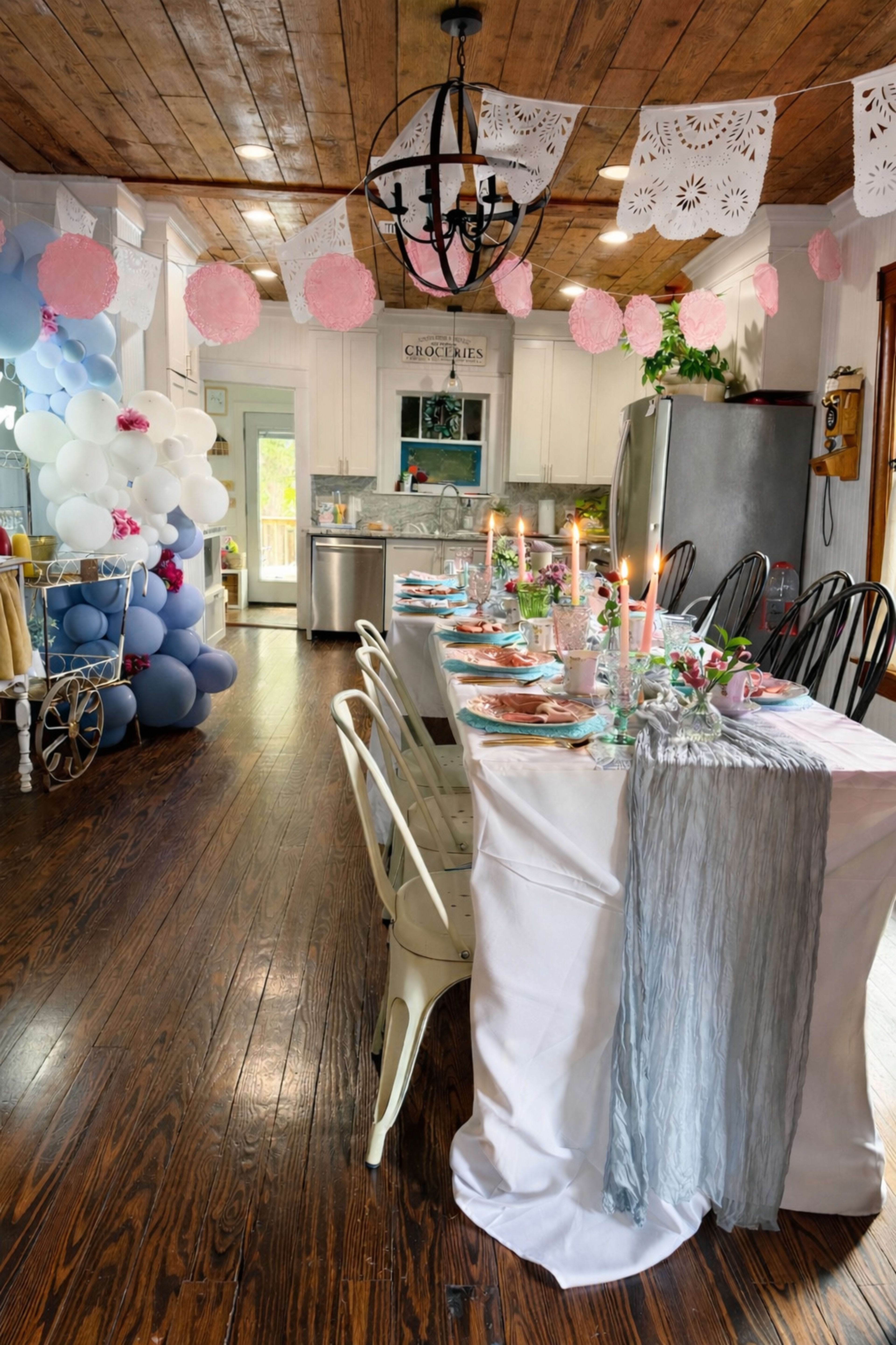 A long dining table is set for a celebration in a rustic kitchen, decorated with colorful balloons, paper bunting, and candles.