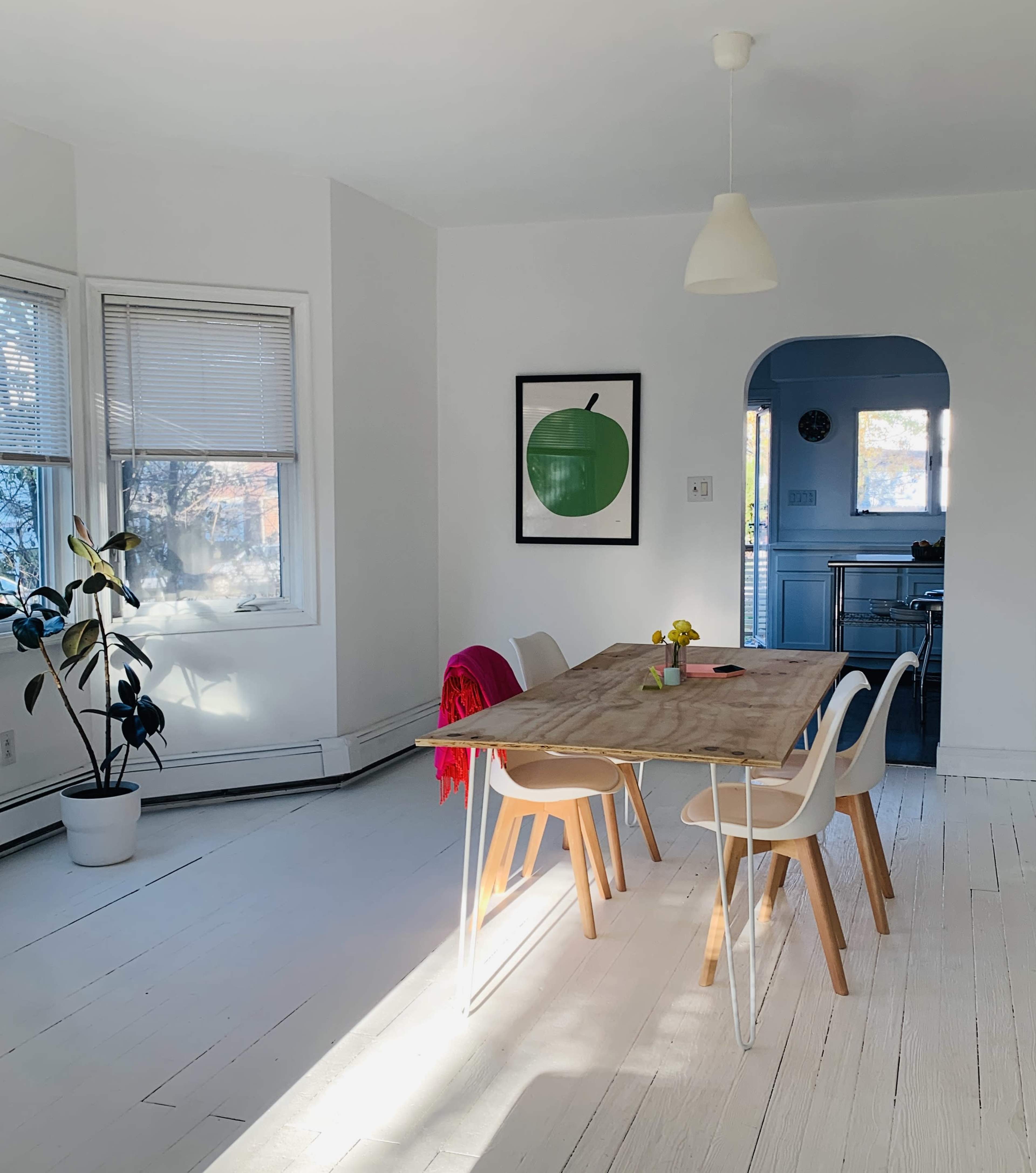 A bright dining area features a wooden table surrounded by white chairs, with a large green apple print on the wall and a potted plant in the corner.