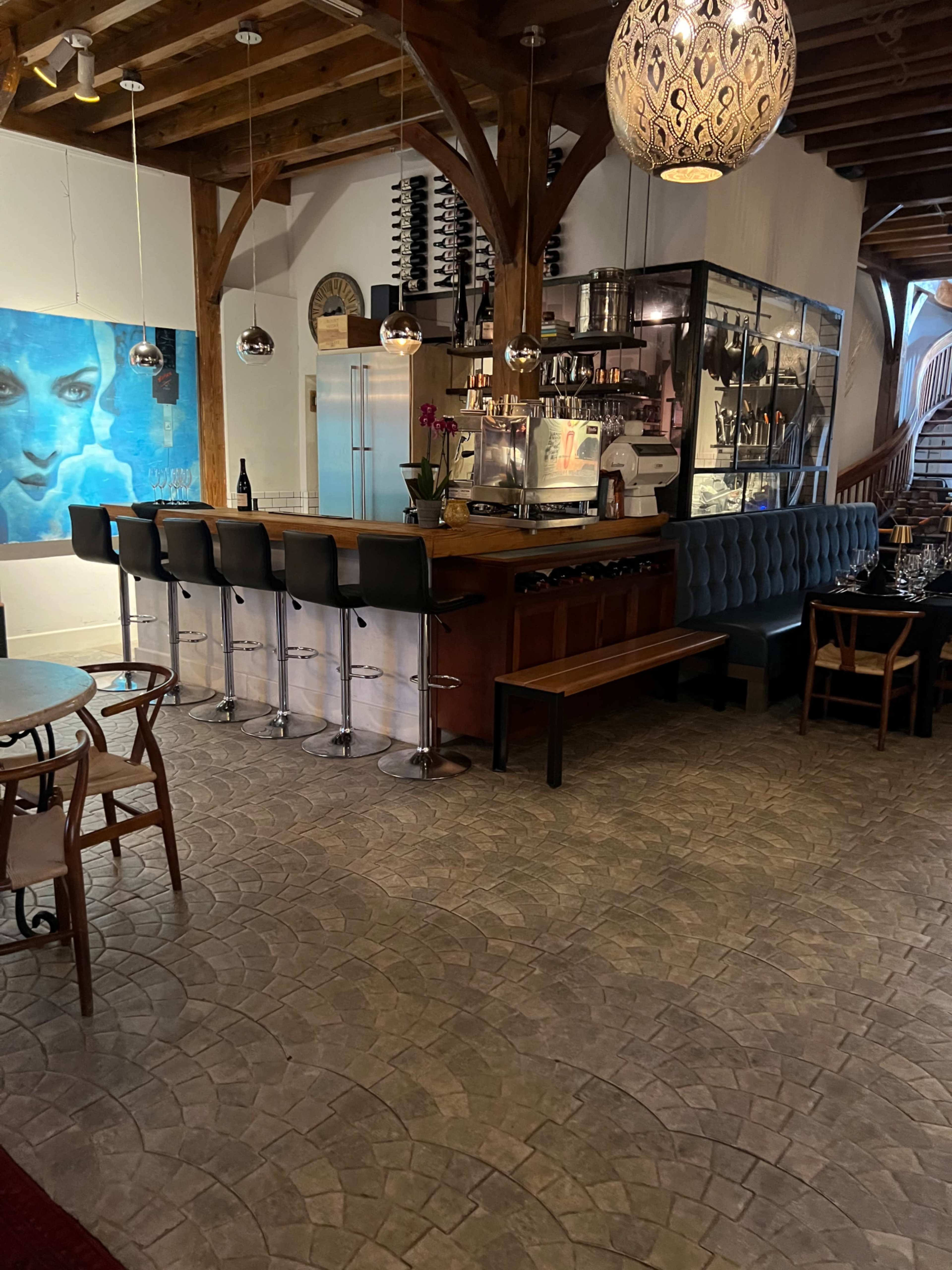 The image shows a cozy restaurant interior featuring a bar with high stools, artistic wall decor, and wooden beams overhead.