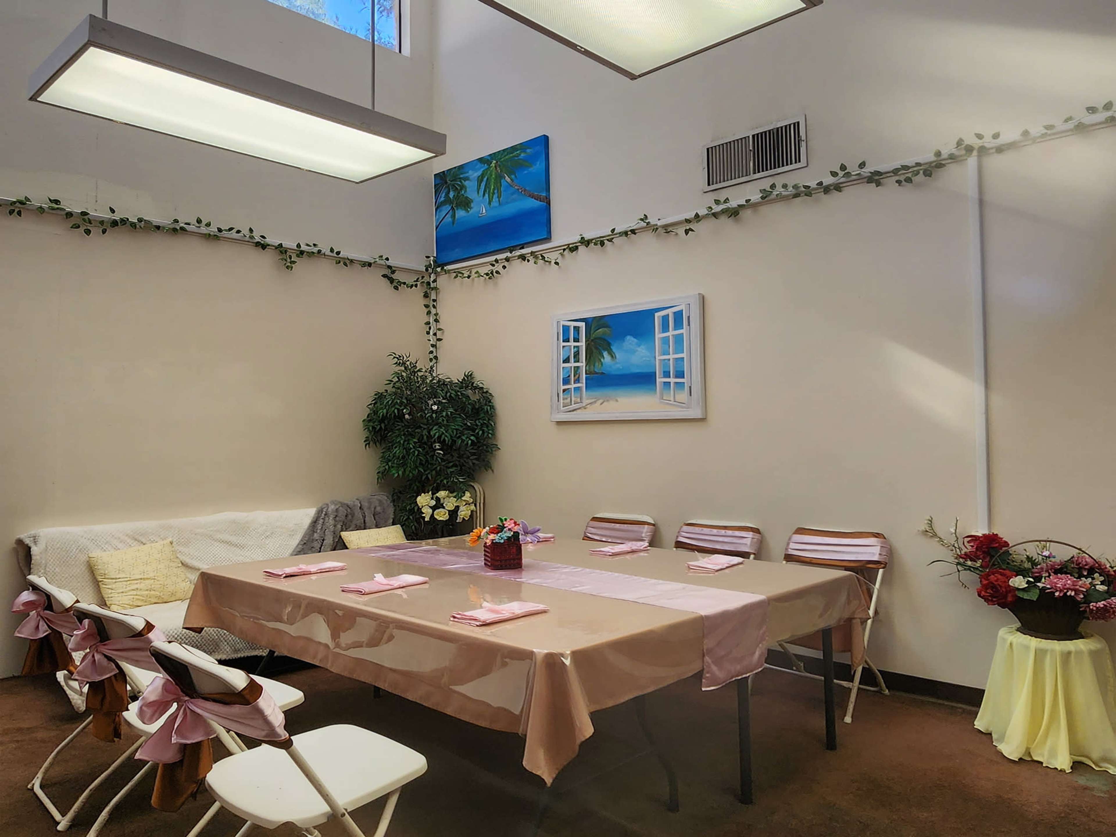The image shows a dining area with a long table set for a meal, surrounded by chairs, and decorated with plants and wall art.