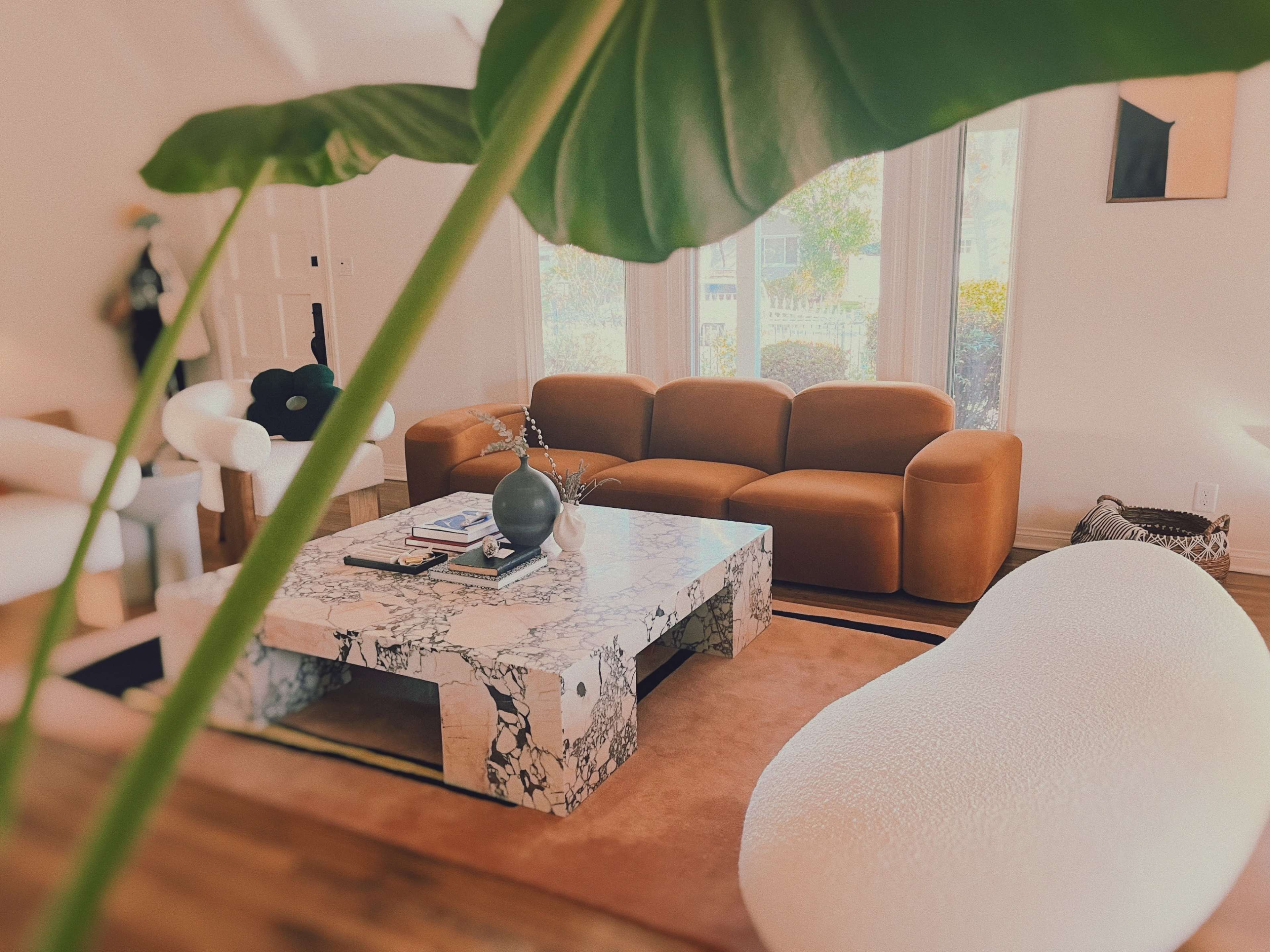 A modern living room featuring a brown sofa, a marble coffee table, and a curved white chair, surrounded by natural light.