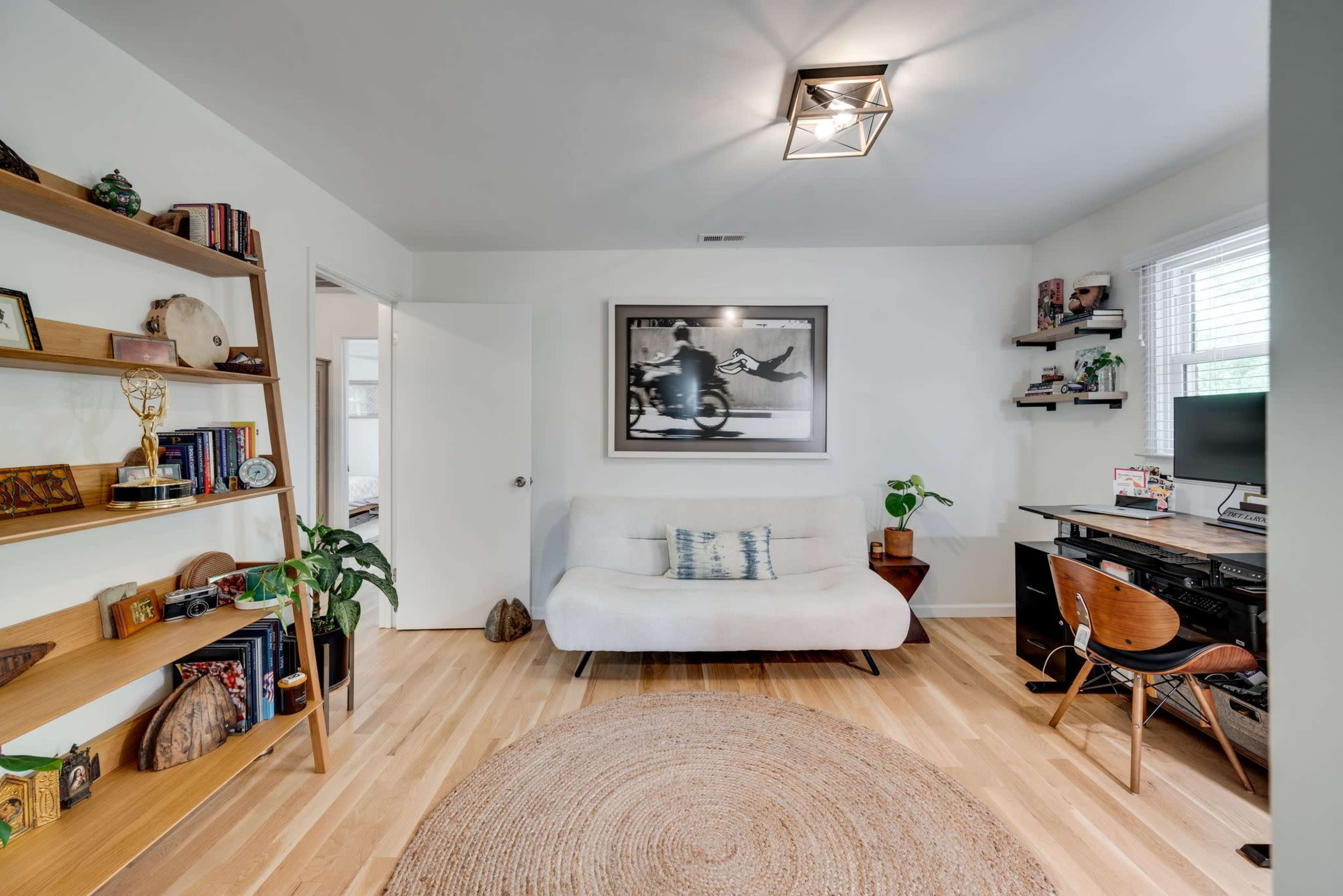 A modern room featuring a light-colored couch, a wooden bookshelf filled with decorative items, a desk with a computer, and a round area rug on the wooden floor.