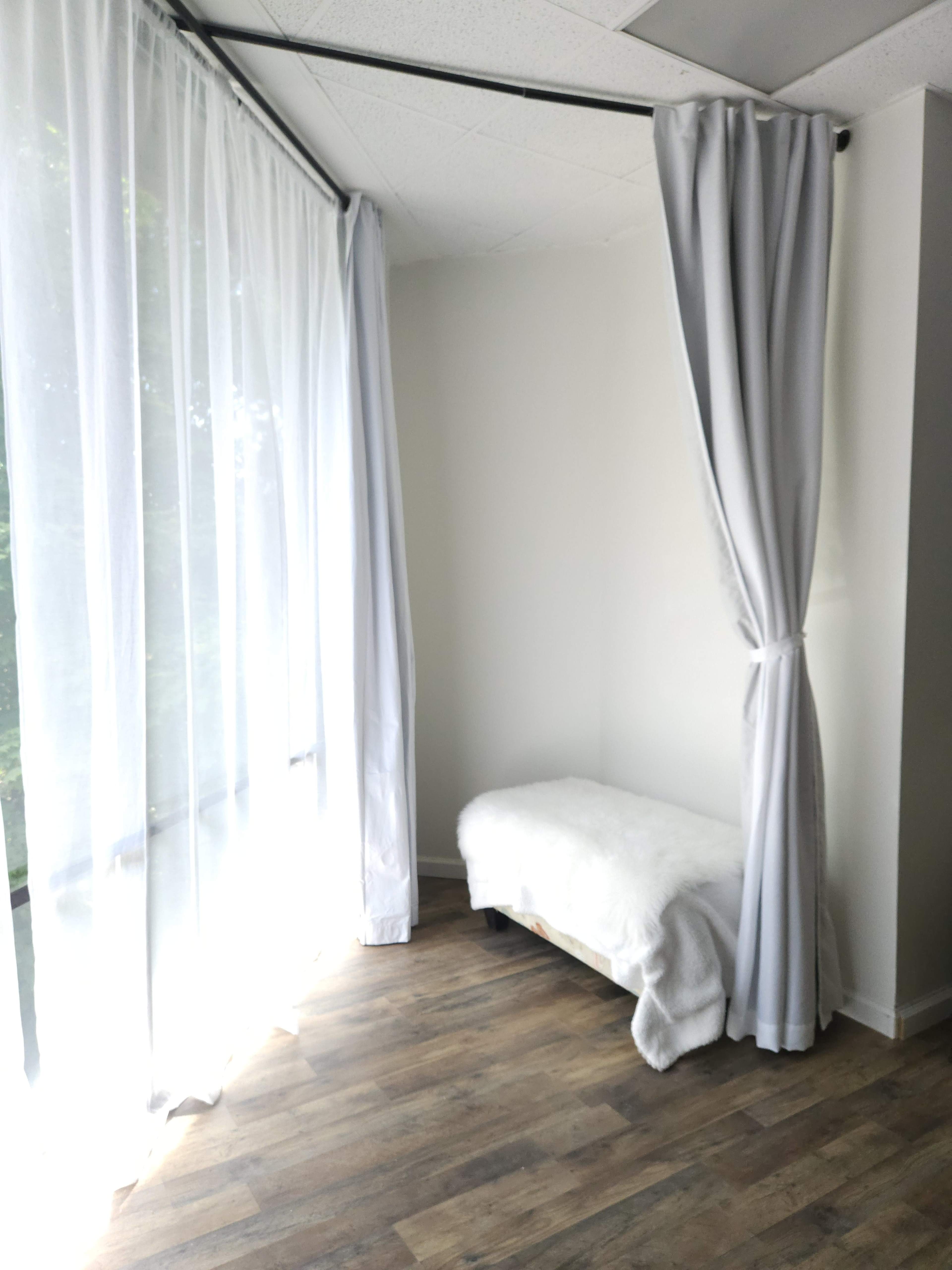 A small, bright room features a bed against a wall with light curtains and large windows allowing natural light.