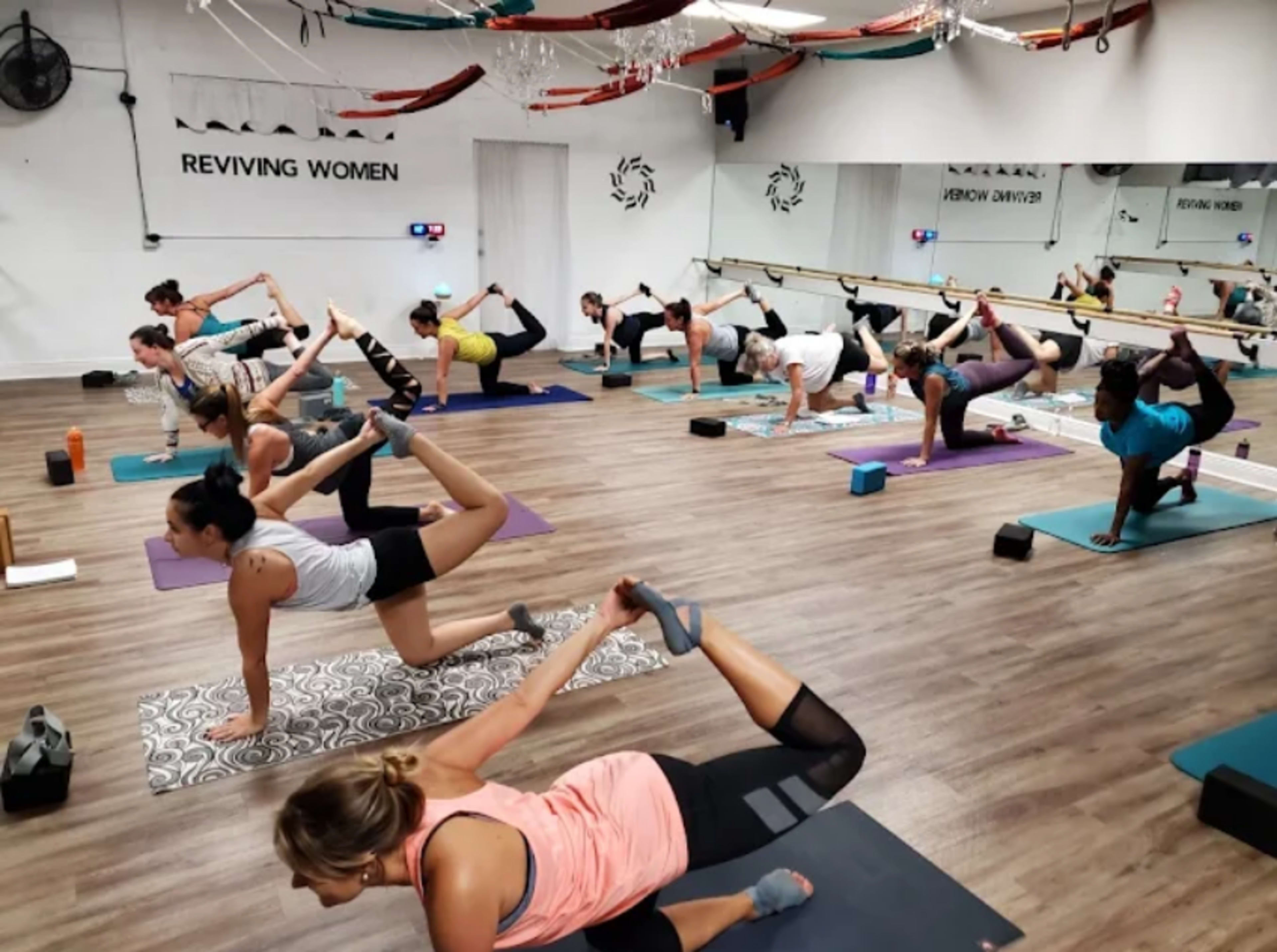 A group of women is engaged in a yoga class, practicing various poses on mats in a brightly lit studio.