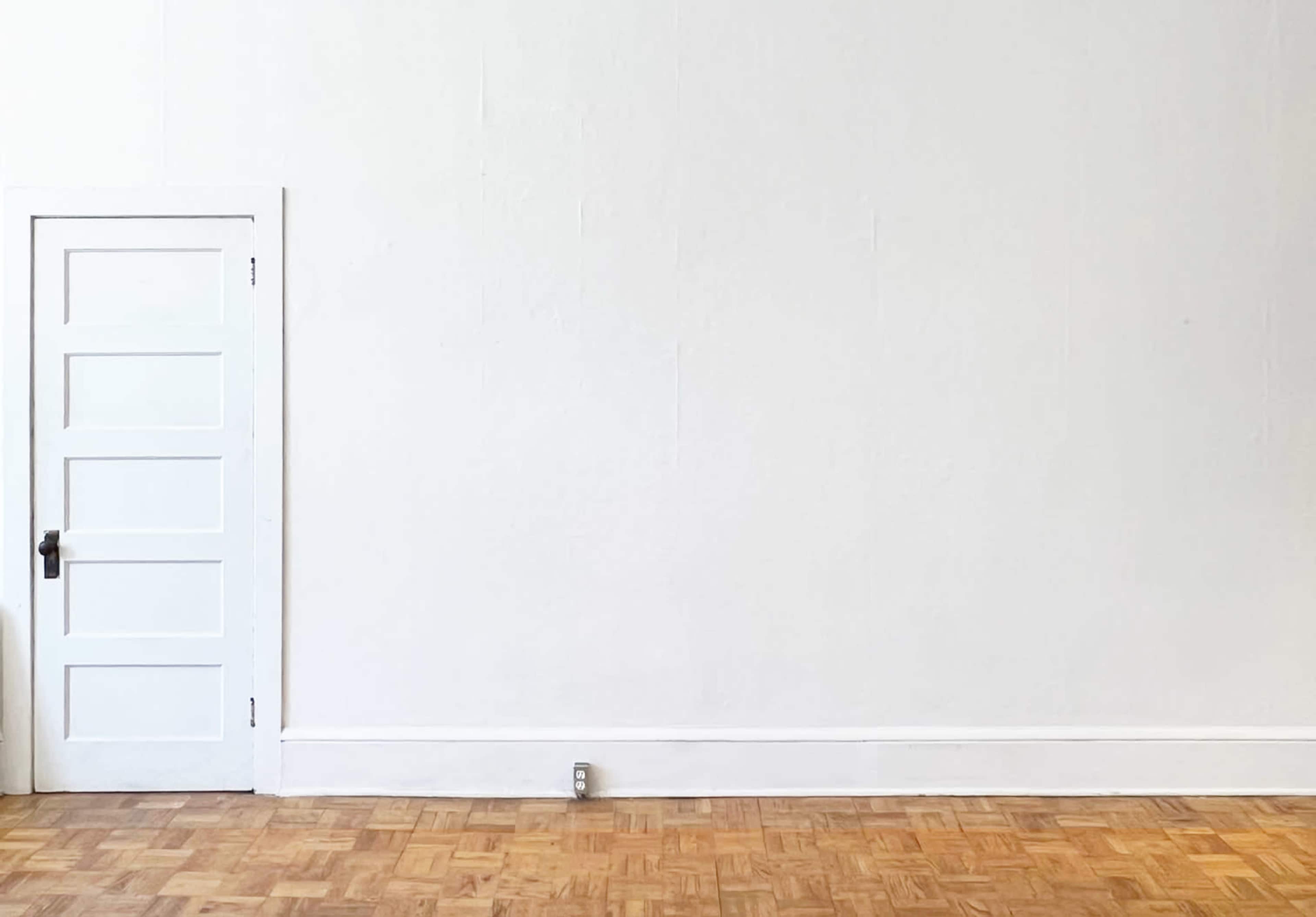 The image shows a simple room with a white wall and a closed door on the left side, adjacent to a wood parquet floor.