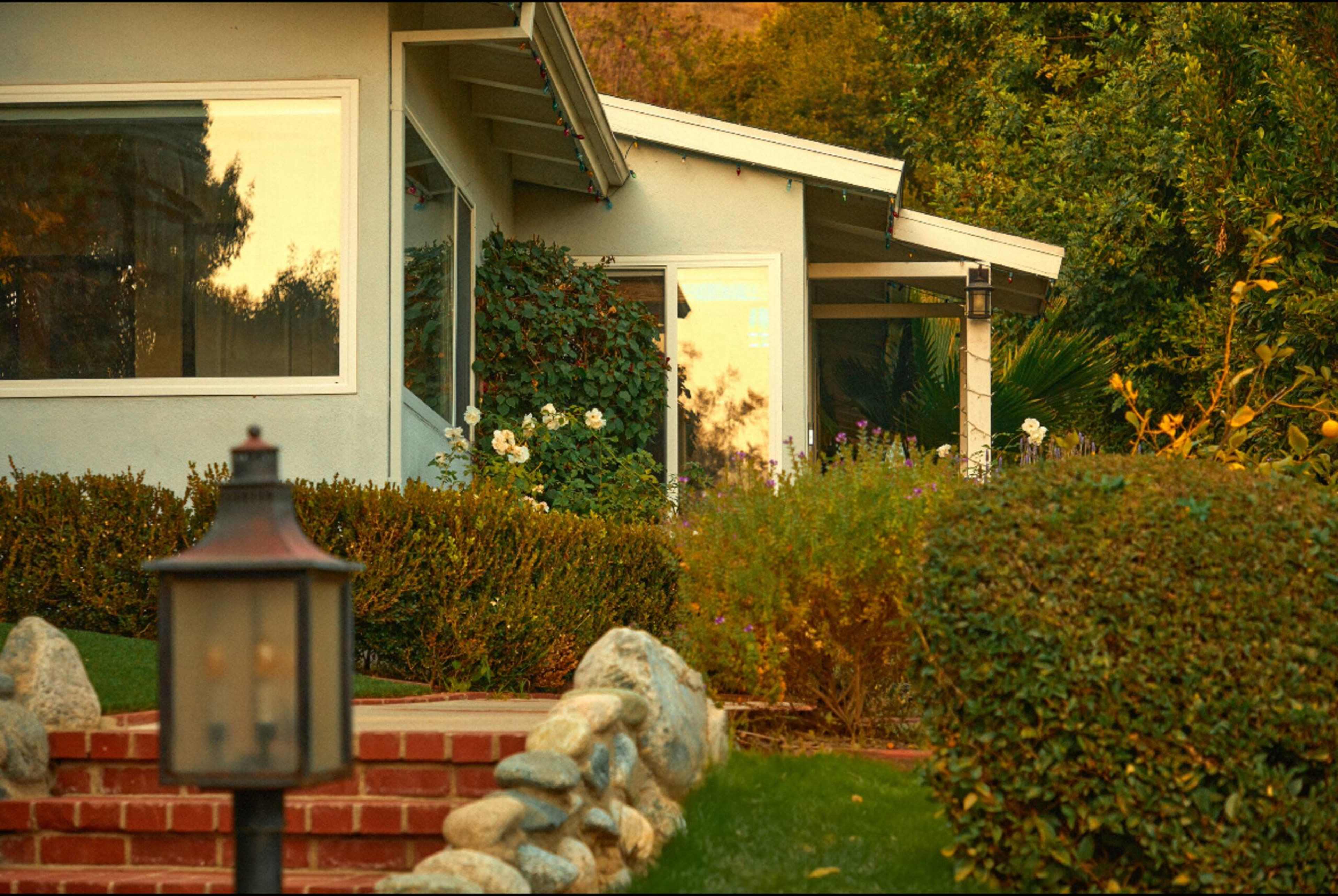 A house with a sloped roof and garden is illuminated by the warm light of sunset, surrounded by neatly trimmed shrubs and flower beds.