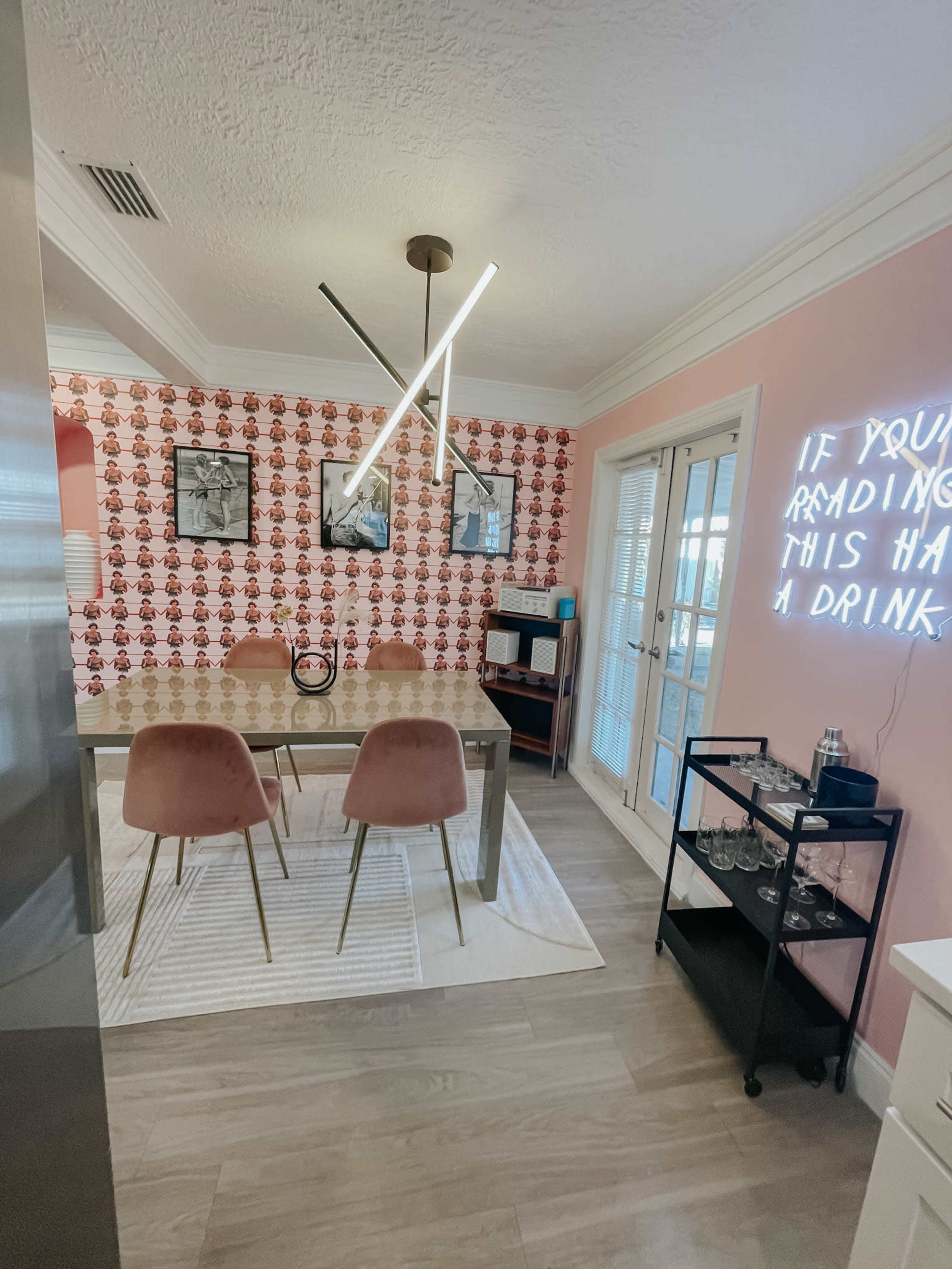 The image shows a dining area with a modern table, pink chairs, patterned wallpaper, and a neon sign on the wall.