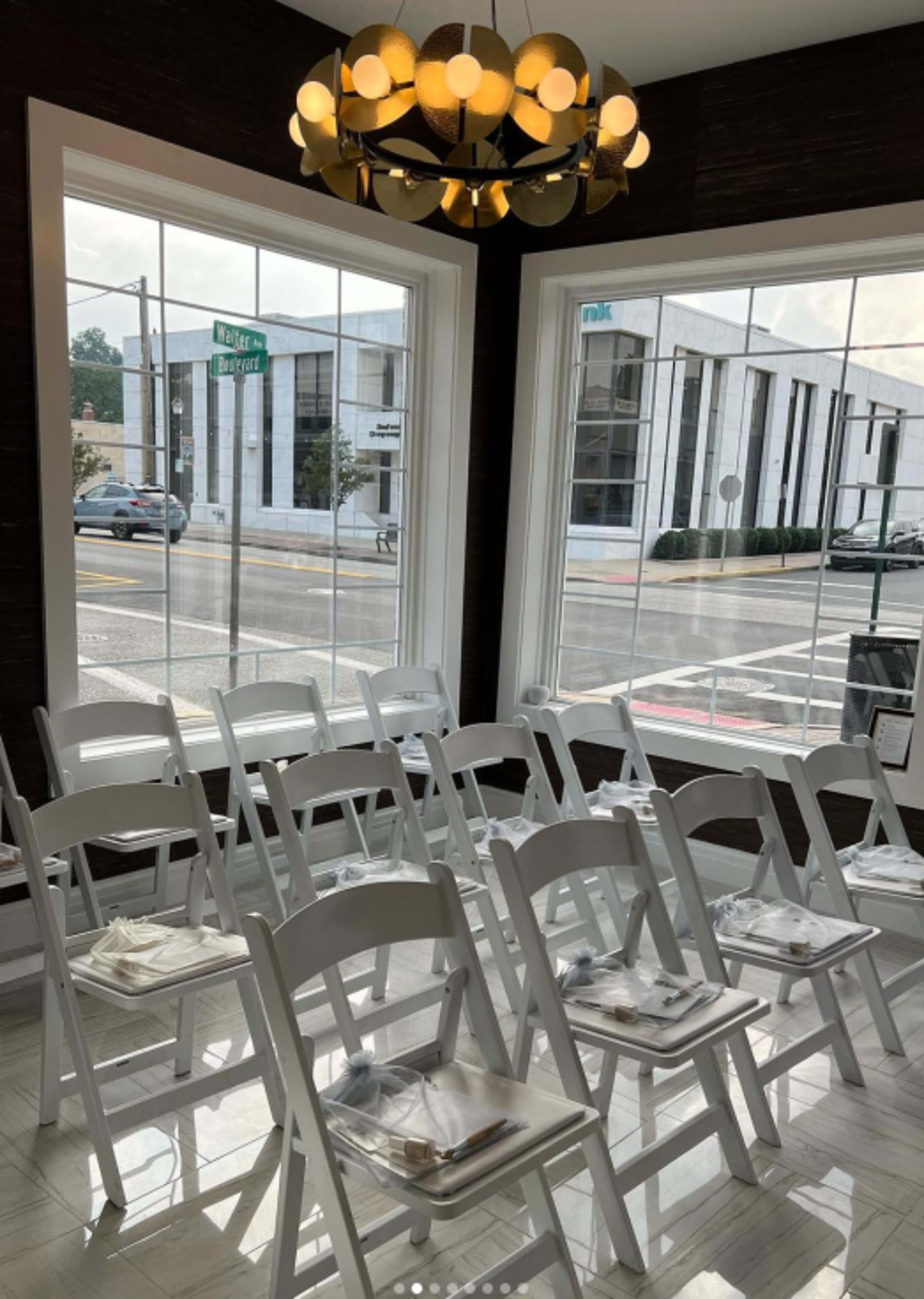 A row of white folding chairs is set up inside a brightly lit room with large windows overlooking a street.