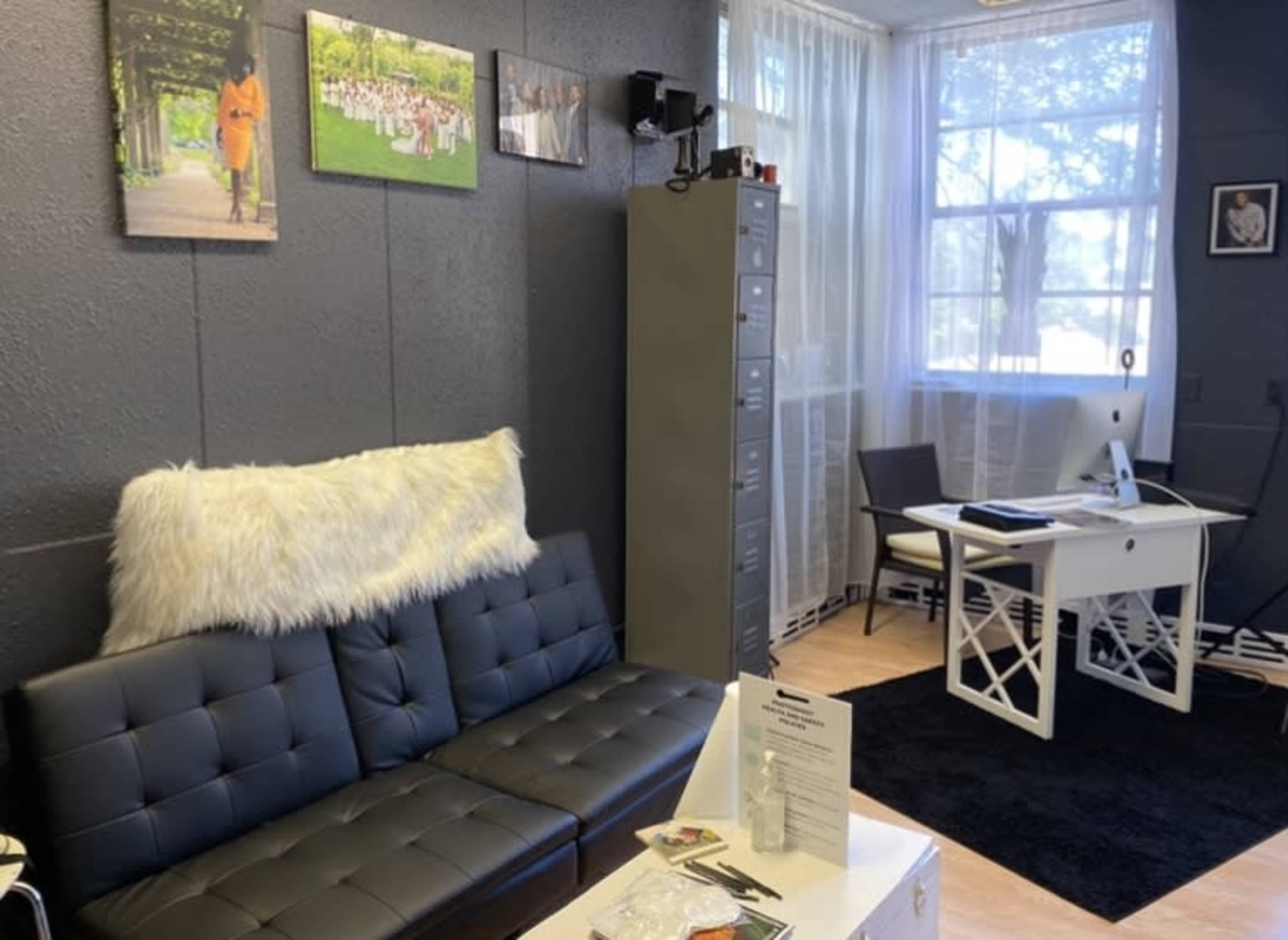 A modern office space featuring a black sofa with a white throw, a desk with a computer, and a window with sheer curtains.