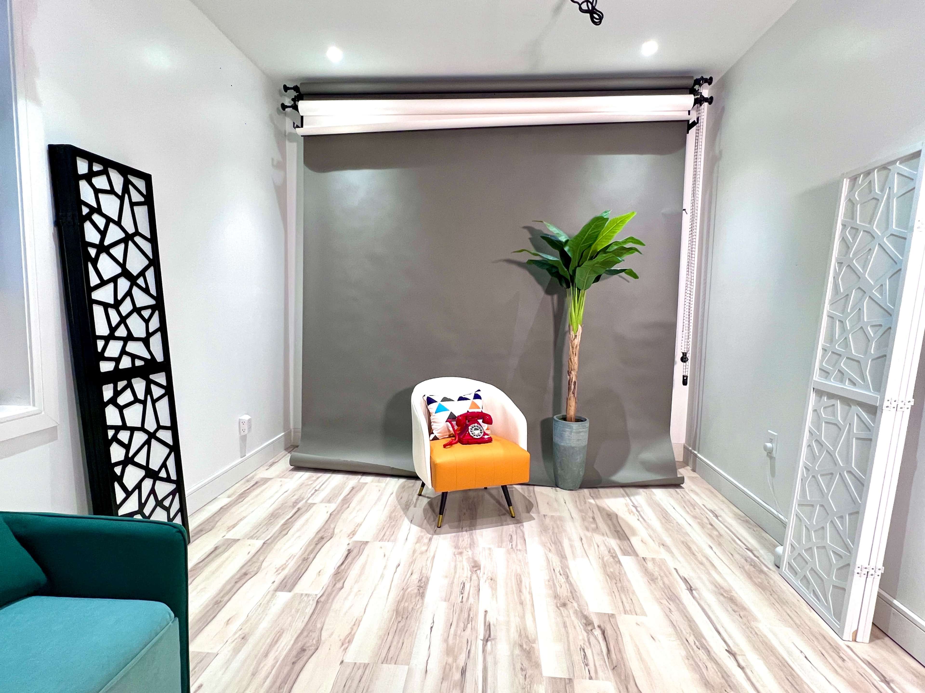 The image shows a minimalist room with a gray backdrop, a bright orange chair, a potted plant, and decorative screens on either side.