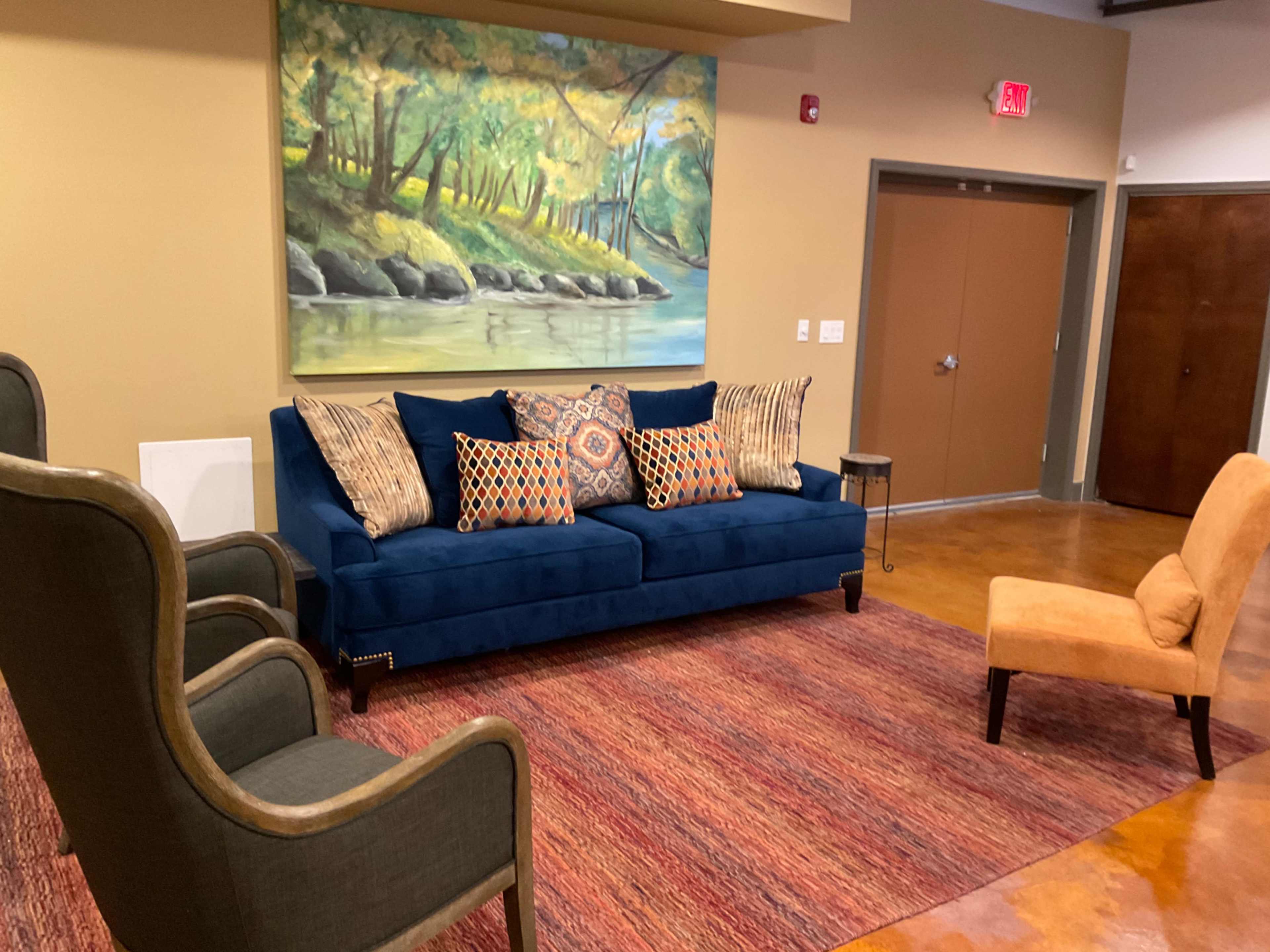 A seating area with a blue sofa adorned with decorative pillows, two armchairs, a small side table, and a painting of a serene landscape on the wall.