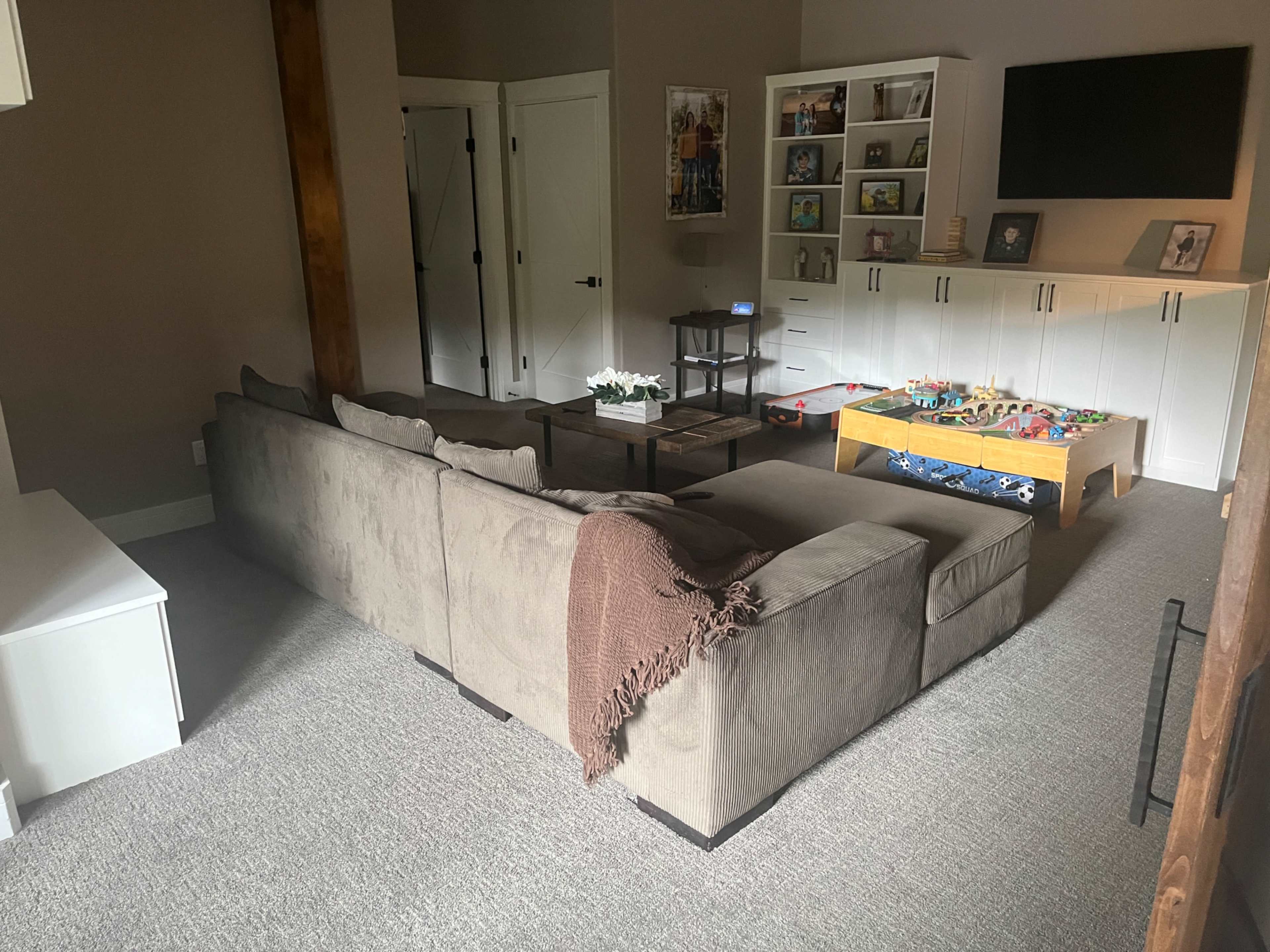 A spacious living room features a large sectional sofa, a coffee table with children's toys, and a TV mounted on the wall above a low storage unit.