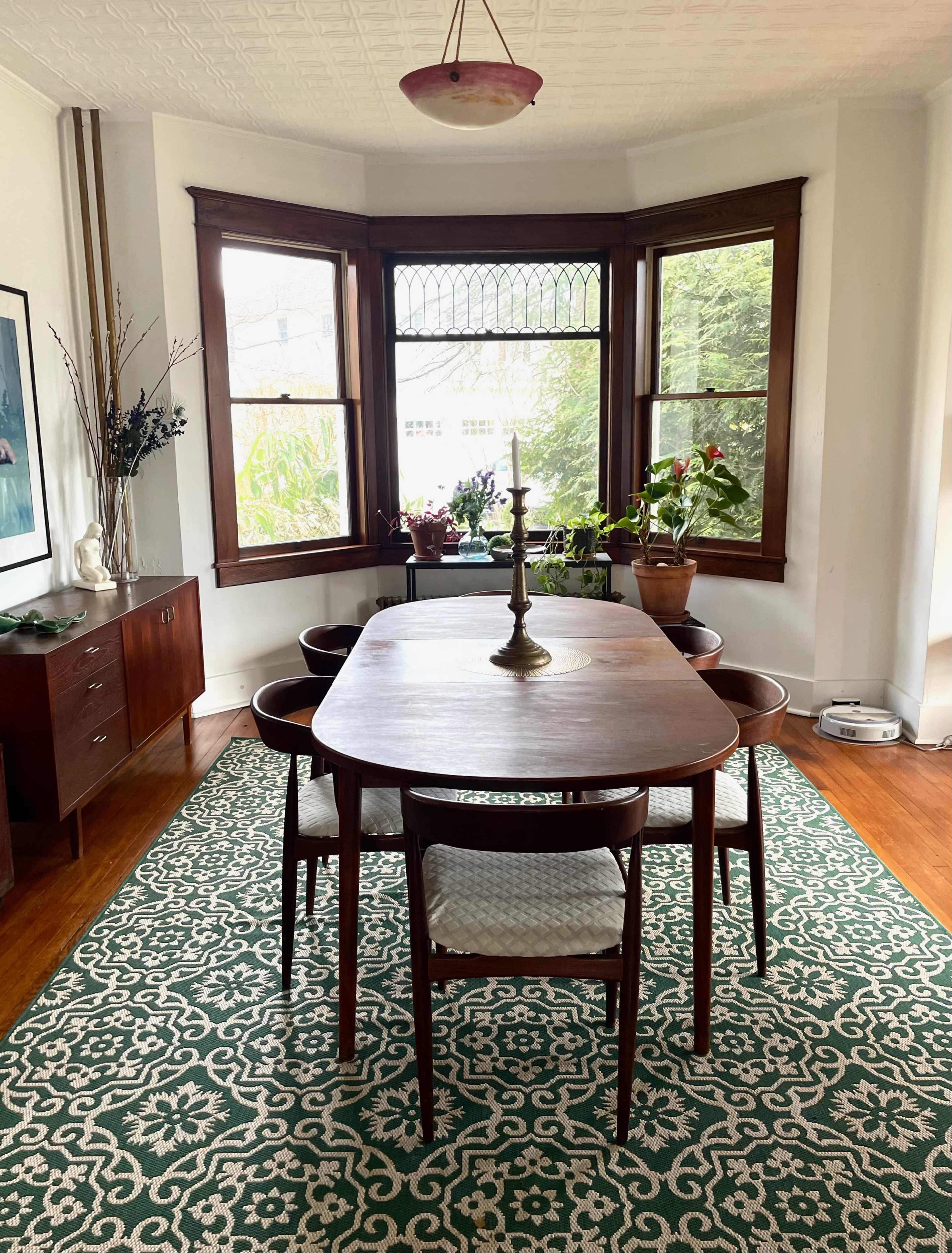 A wooden dining table with chairs is set in a bay window area surrounded by plants and a sideboard.