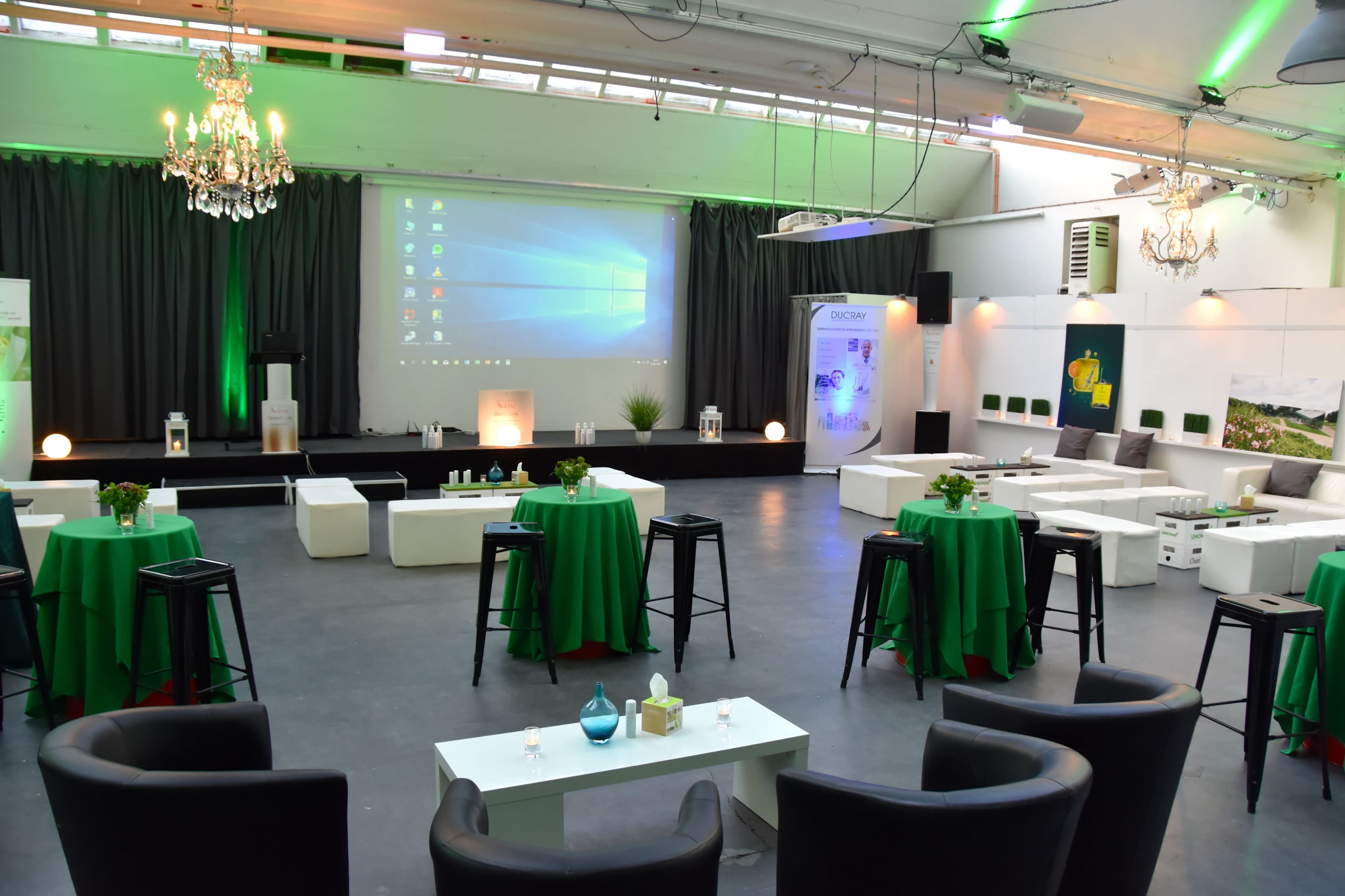 A spacious event venue set up with green tables and black stools, featuring a presentation screen and decorative plants.