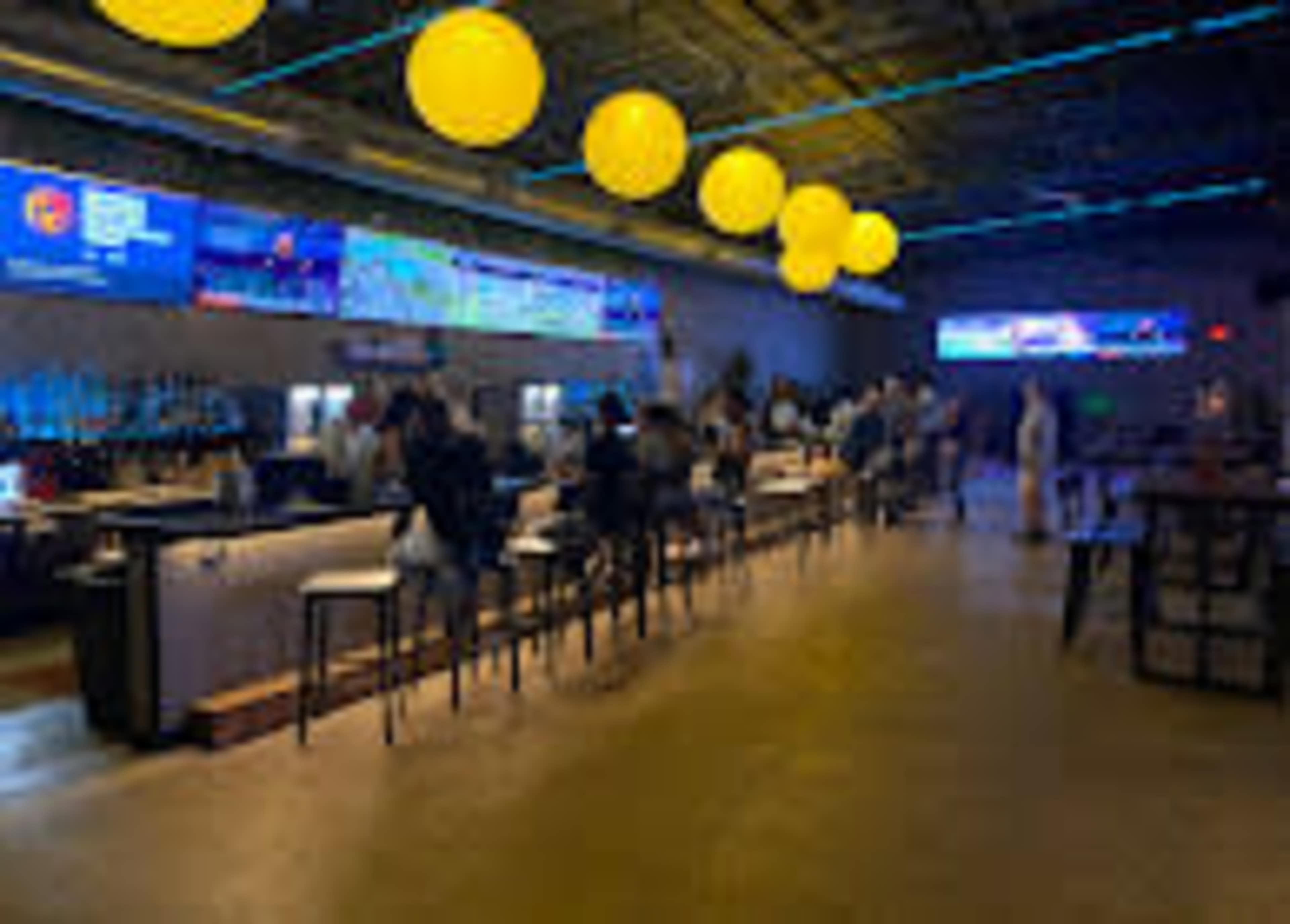 The image shows a modern bar area with patrons seated at the counter and bright yellow pendant lights hanging from the ceiling.