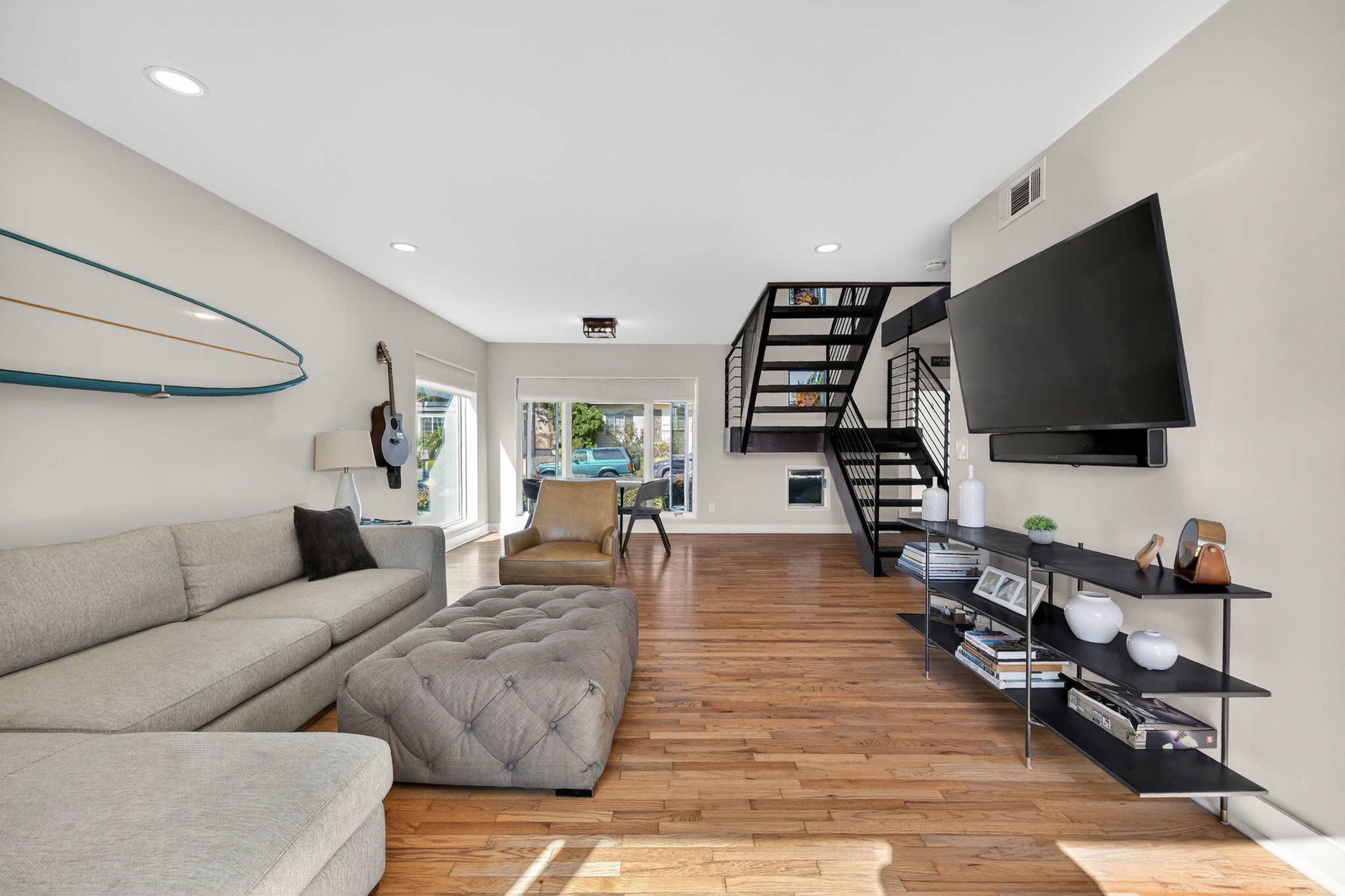 The image shows a modern living room with a sectional sofa, a tufted ottoman, a wall-mounted television, and a staircase in the background.