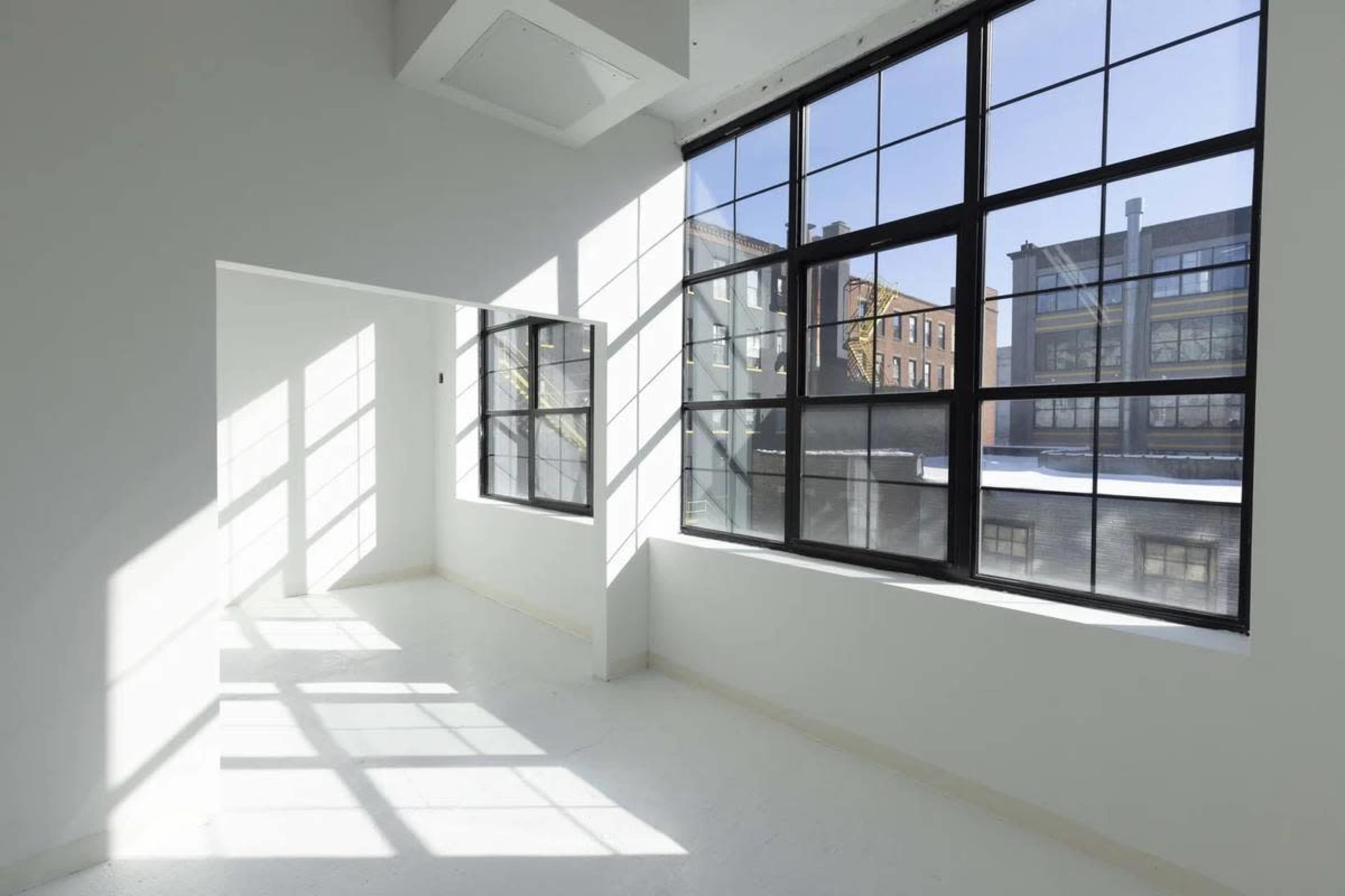 The image shows a bright, empty room with large black-framed windows casting shadows on the white walls and floor.