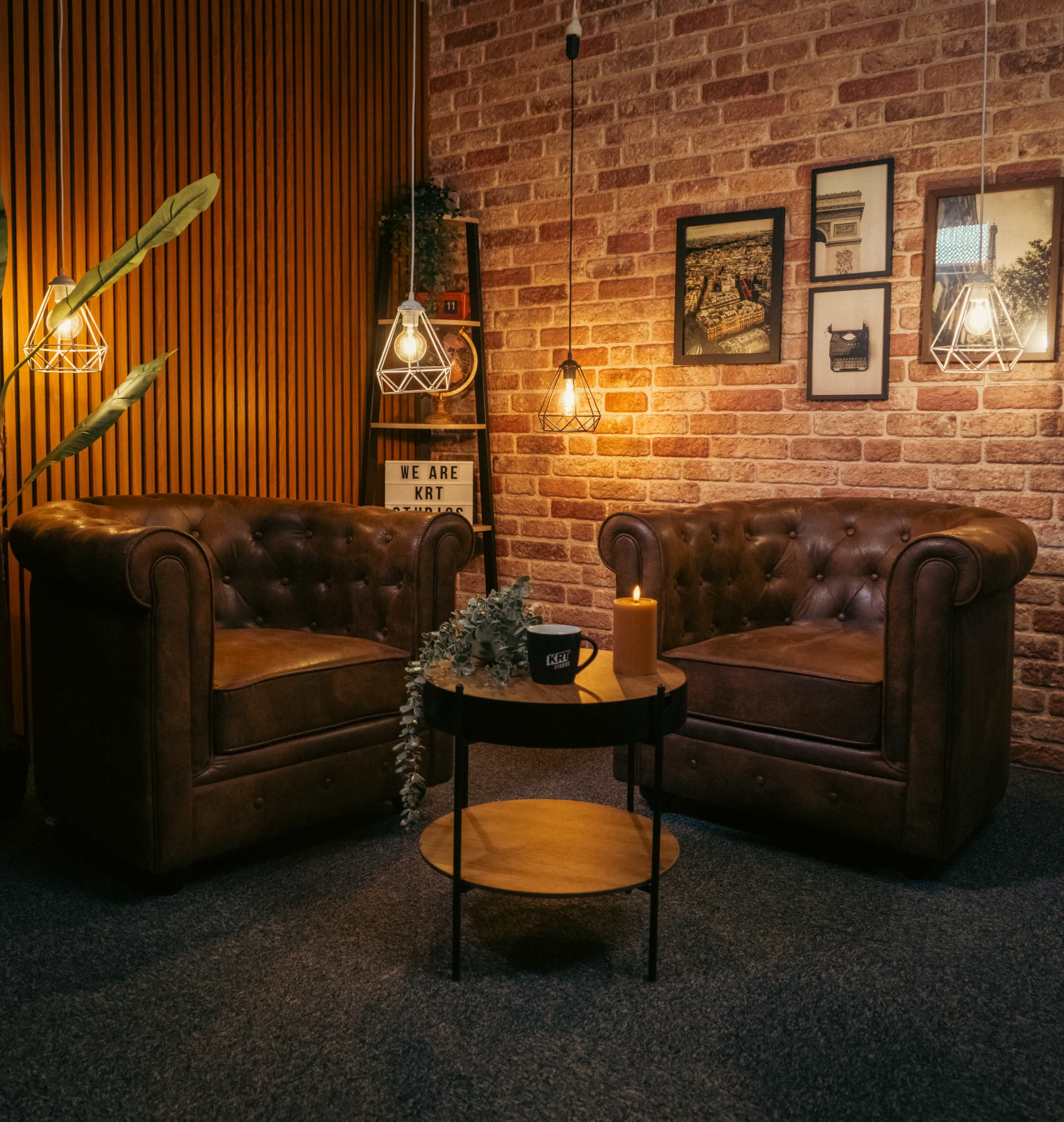 The image shows two brown leather chesterfield-style armchairs facing a small round table with a candle and plants, surrounded by warm lighting and decorative wall frames.