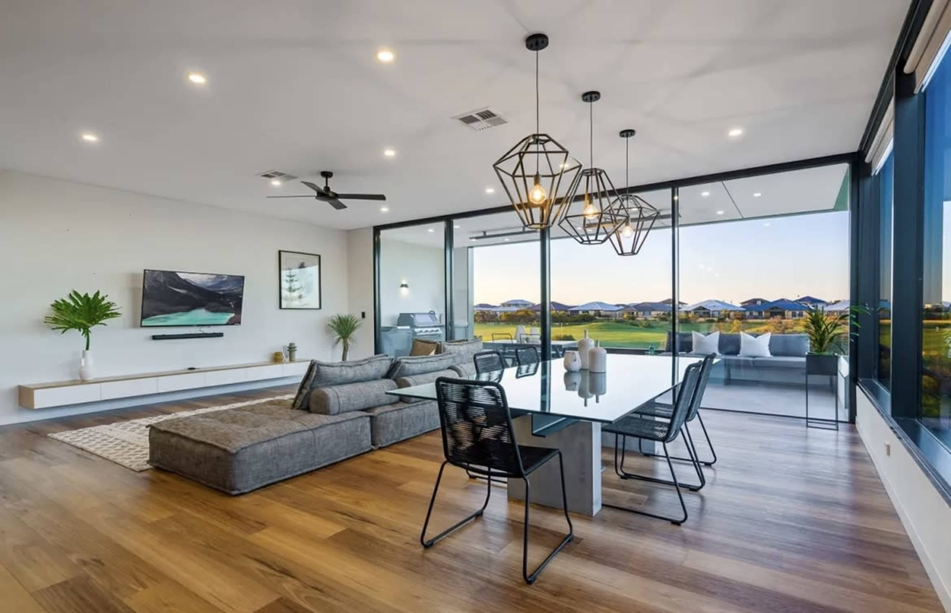 The image shows a modern living room with a large sectional sofa, a dining table, and expansive windows showcasing a scenic view of hills in the background.