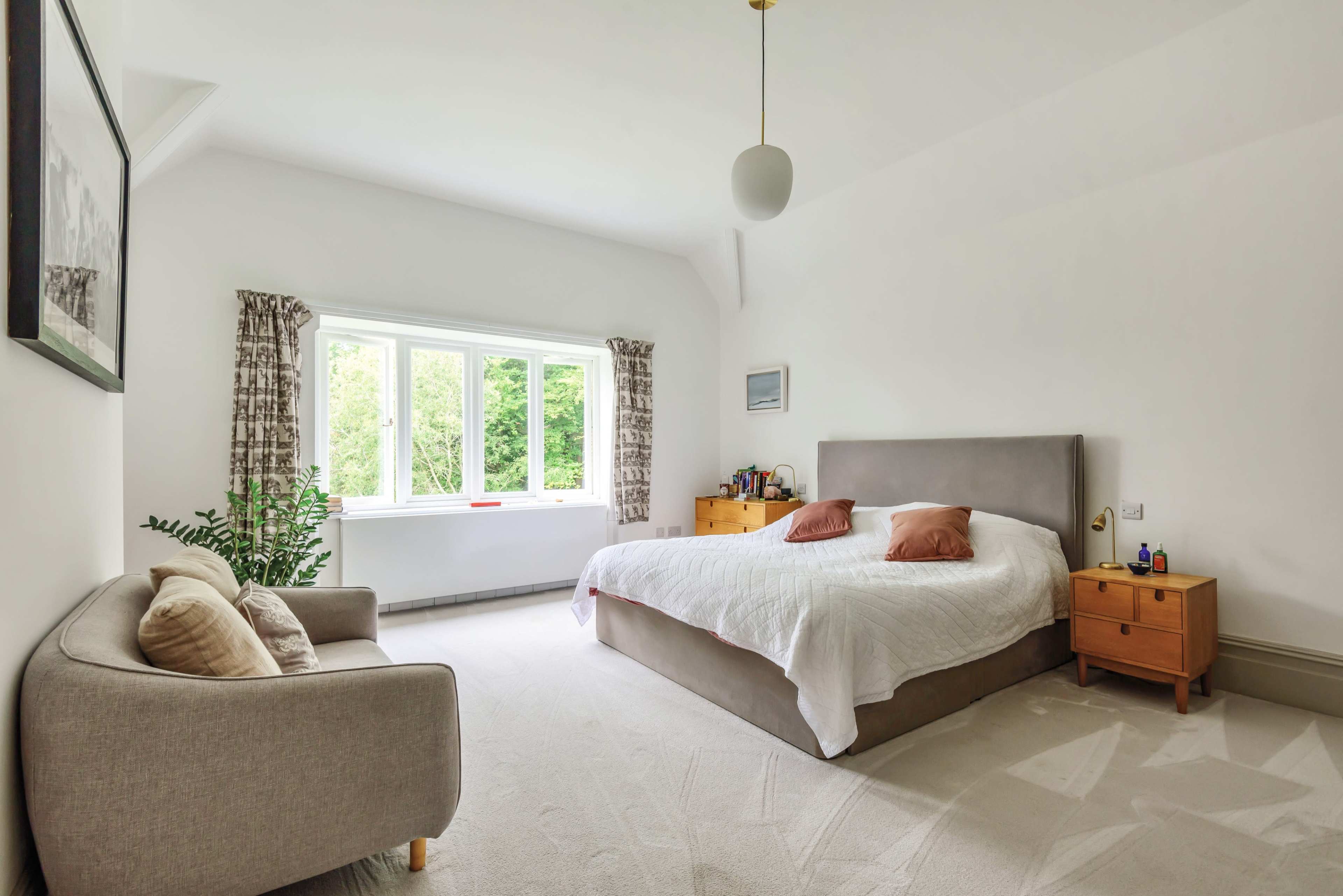 A bright bedroom features a large bed with a gray headboard, flanked by two wooden nightstands, a couch, and a large window overlooking greenery.