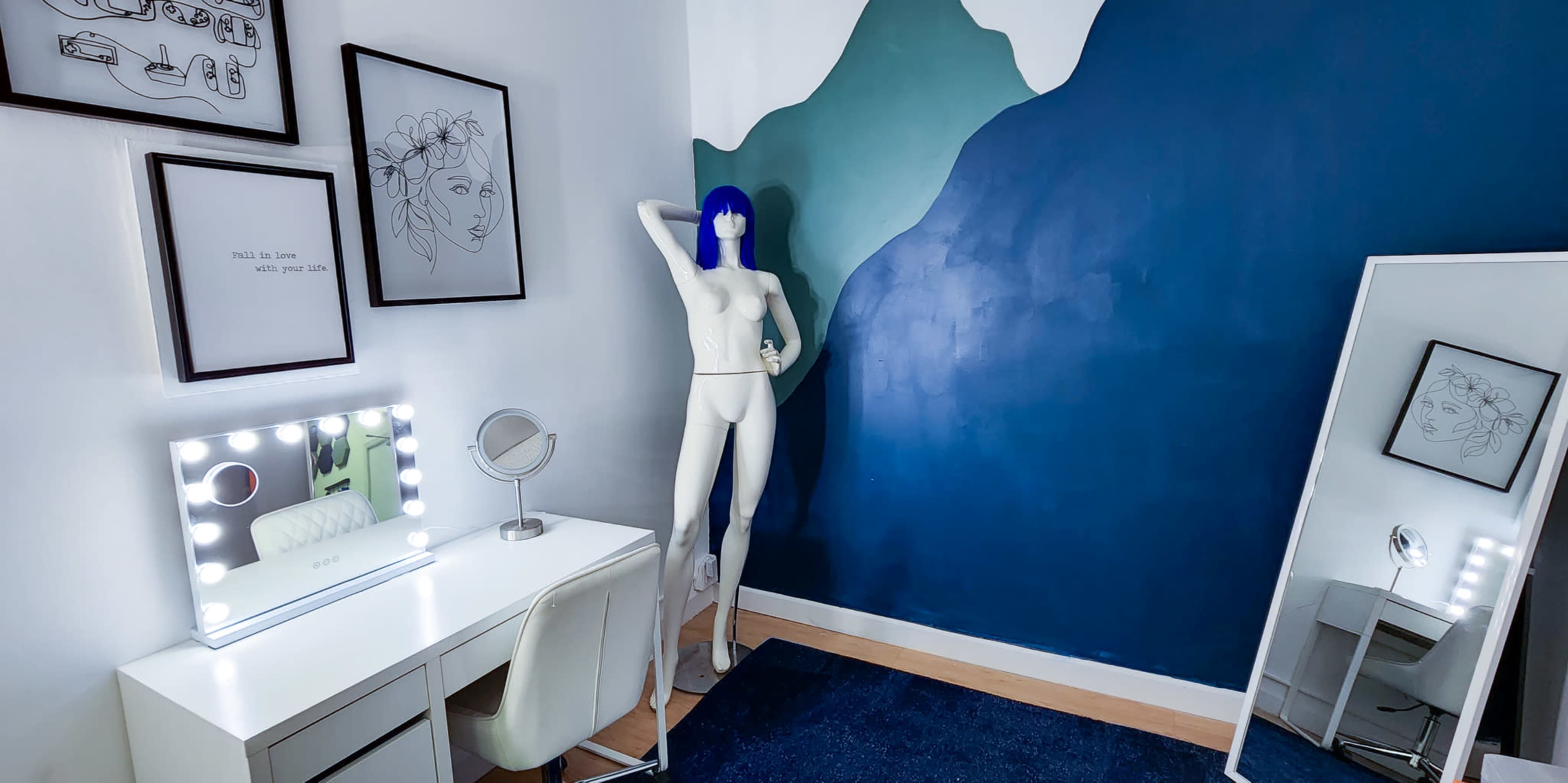 A room featuring a white vanity desk with a mirror, a mannequin with blue hair, and a wall painted in a gradient of green and blue.