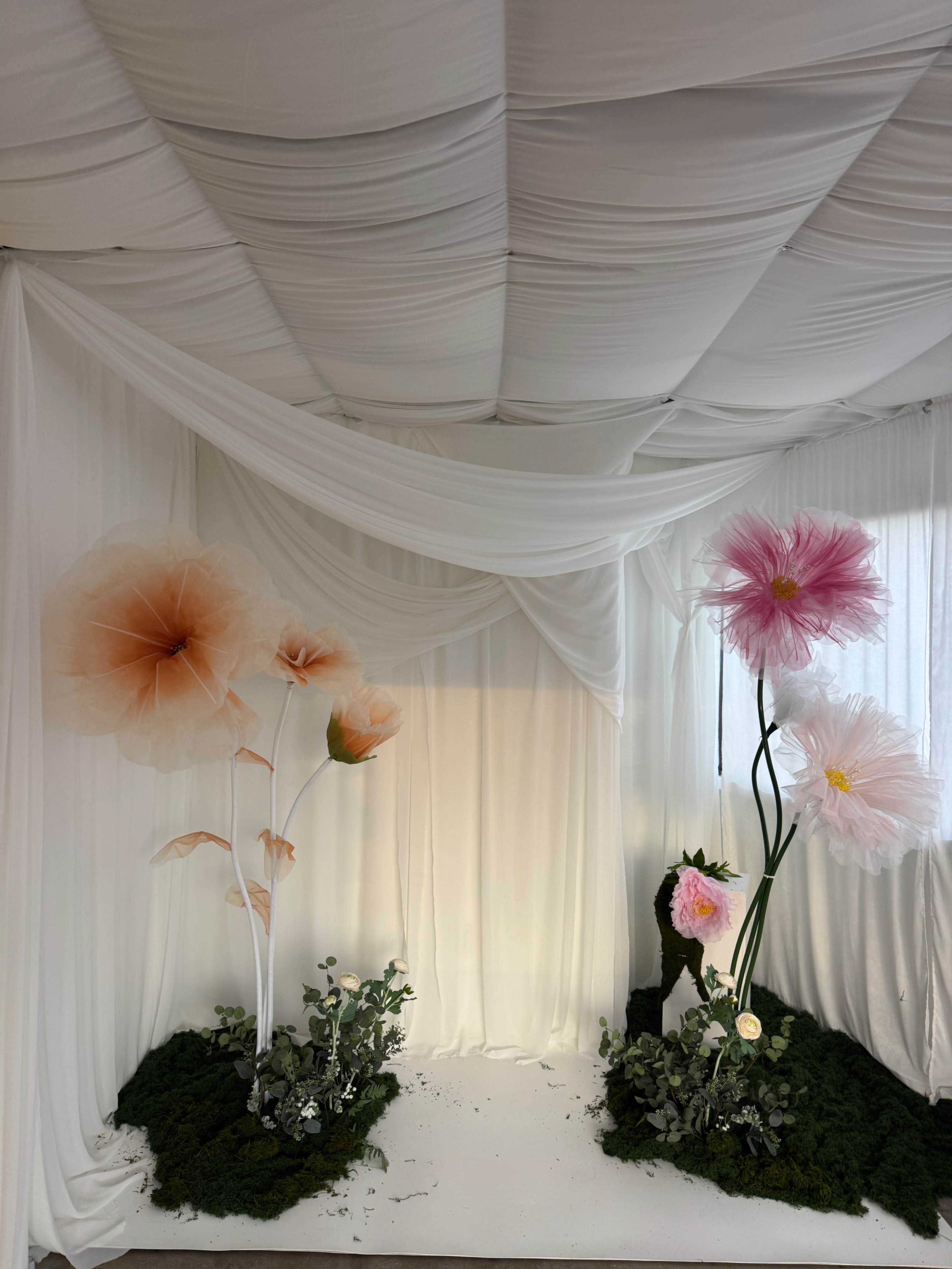 The image depicts a decorative scene featuring oversized artificial flowers arranged on either side of a white draped backdrop, with lush greenery at the base.