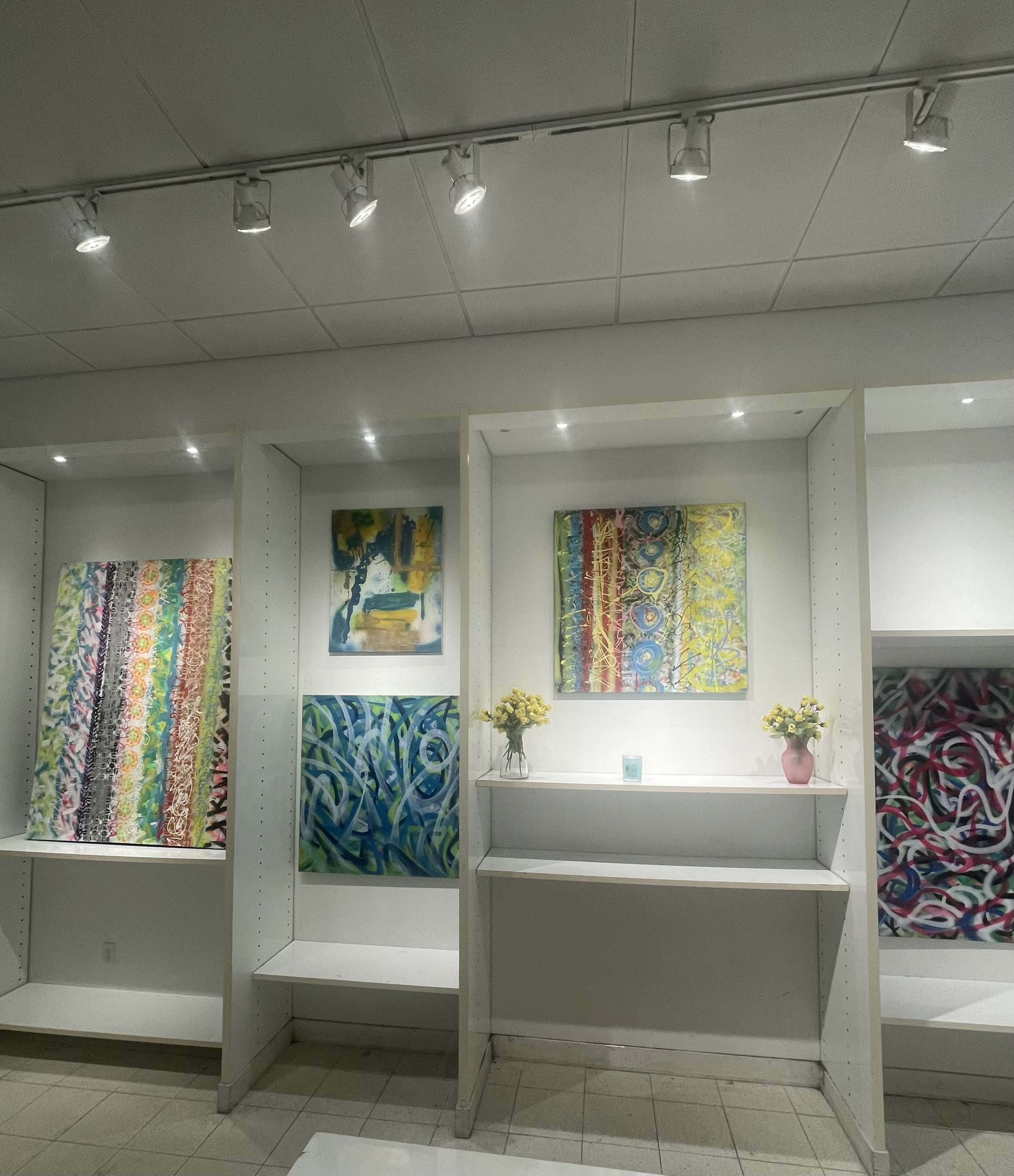 The image shows an art gallery featuring colorful abstract paintings displayed on white shelves with flowers on a small platform.