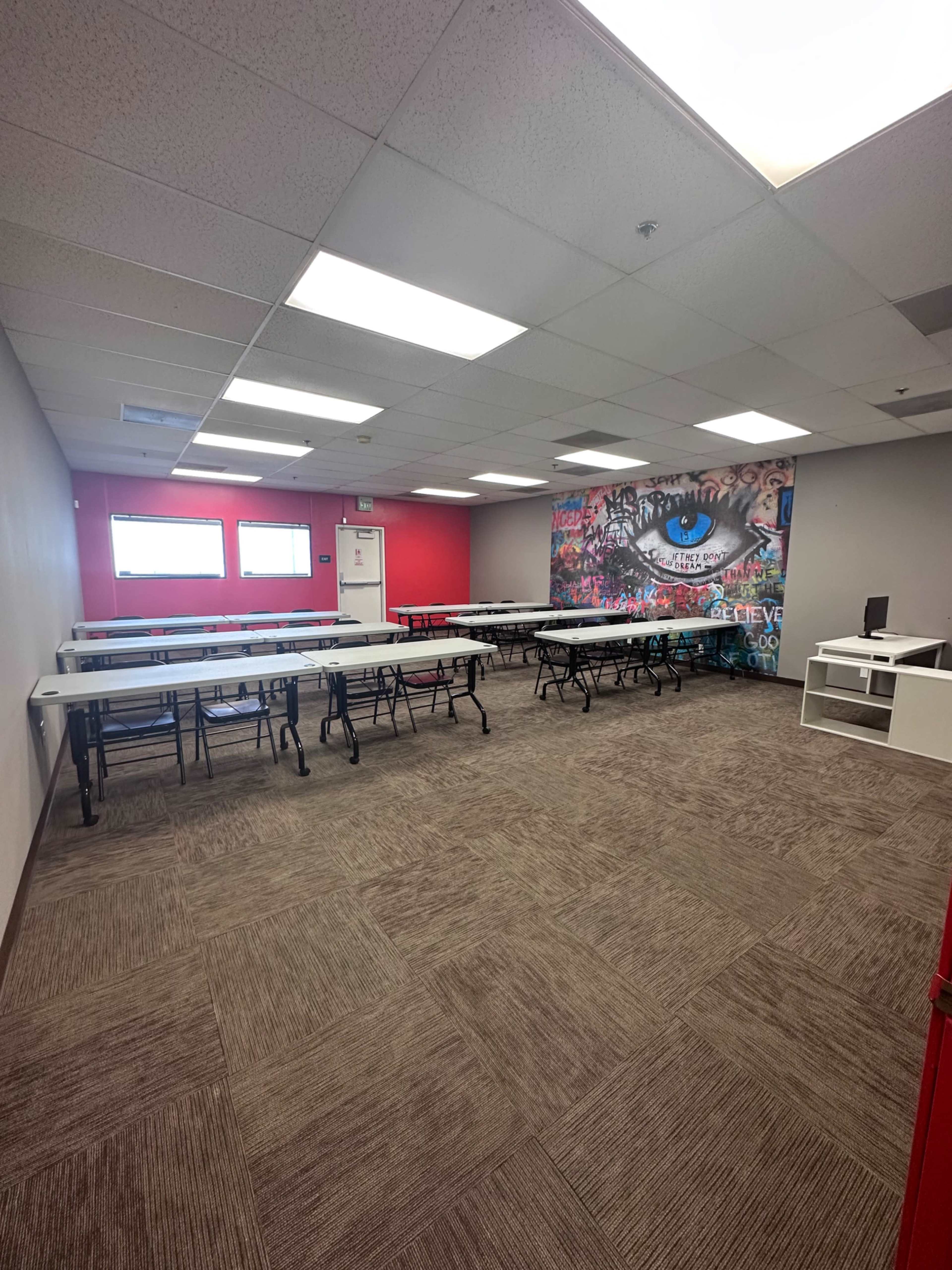 A spacious room filled with folding tables arranged in rows, featuring a colorful mural on one wall and large windows allowing natural light.