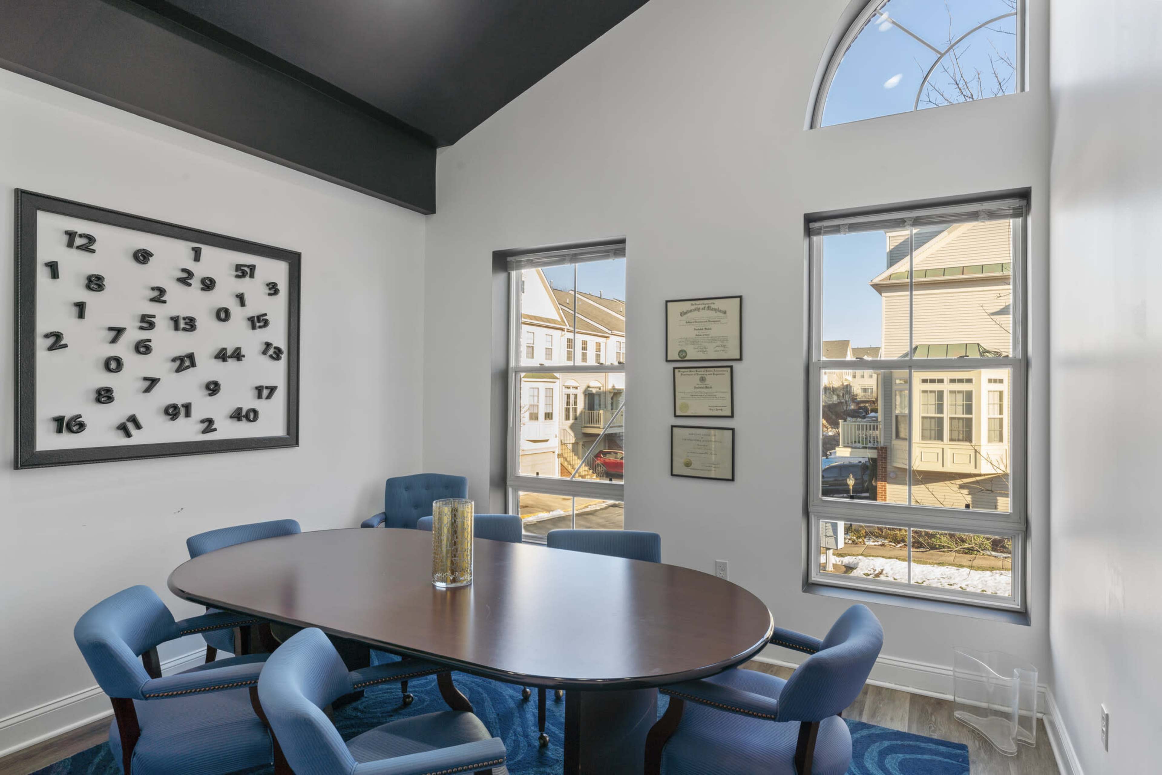 A conference room is set up with a large oval table surrounded by blue upholstered chairs, and features a large clock on the wall and certificates displayed in frames.