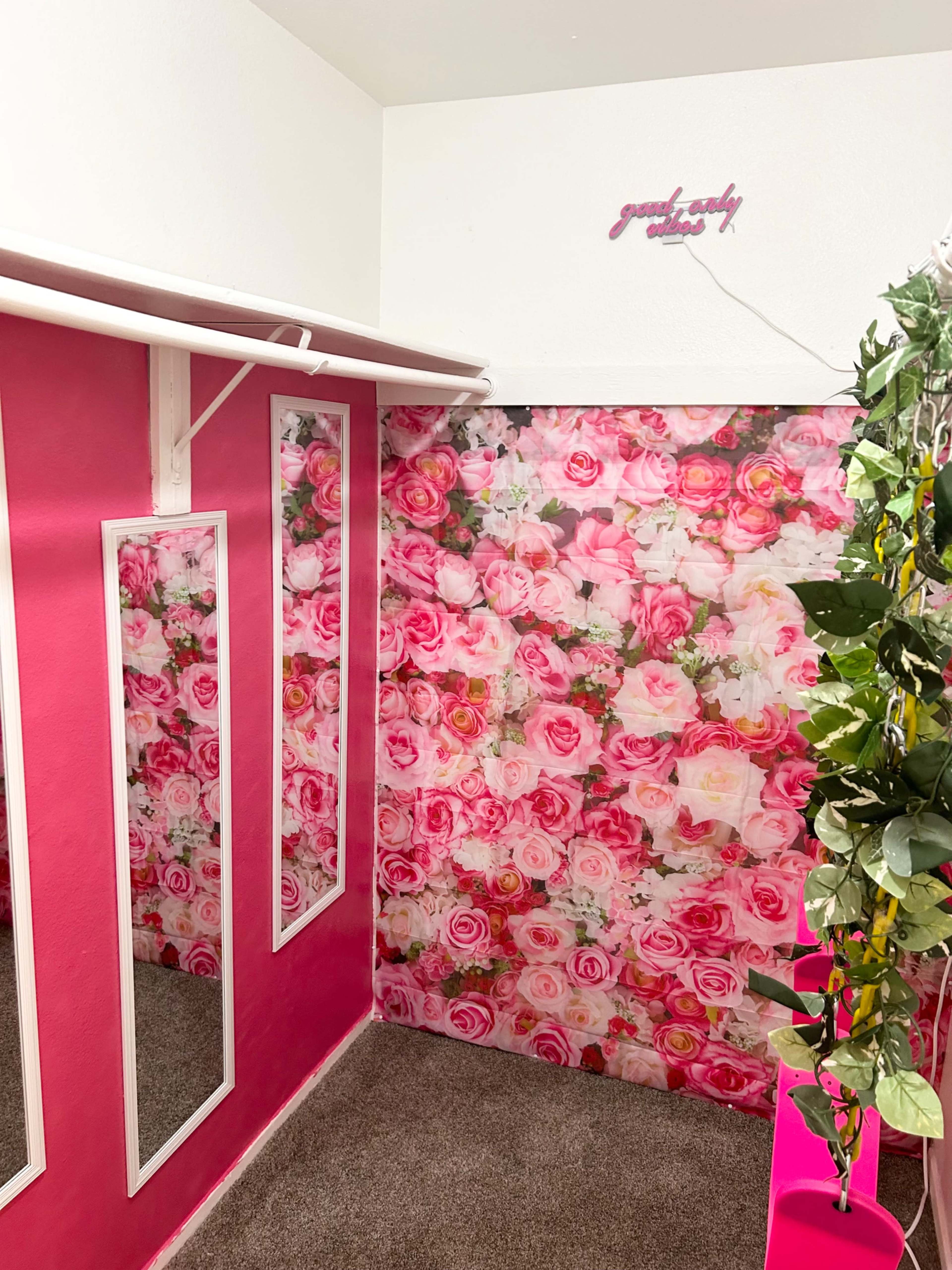 The image shows a corner of a room decorated with a floral wallpaper featuring pink roses, alongside mirrors on one wall and a green plant hanging nearby.