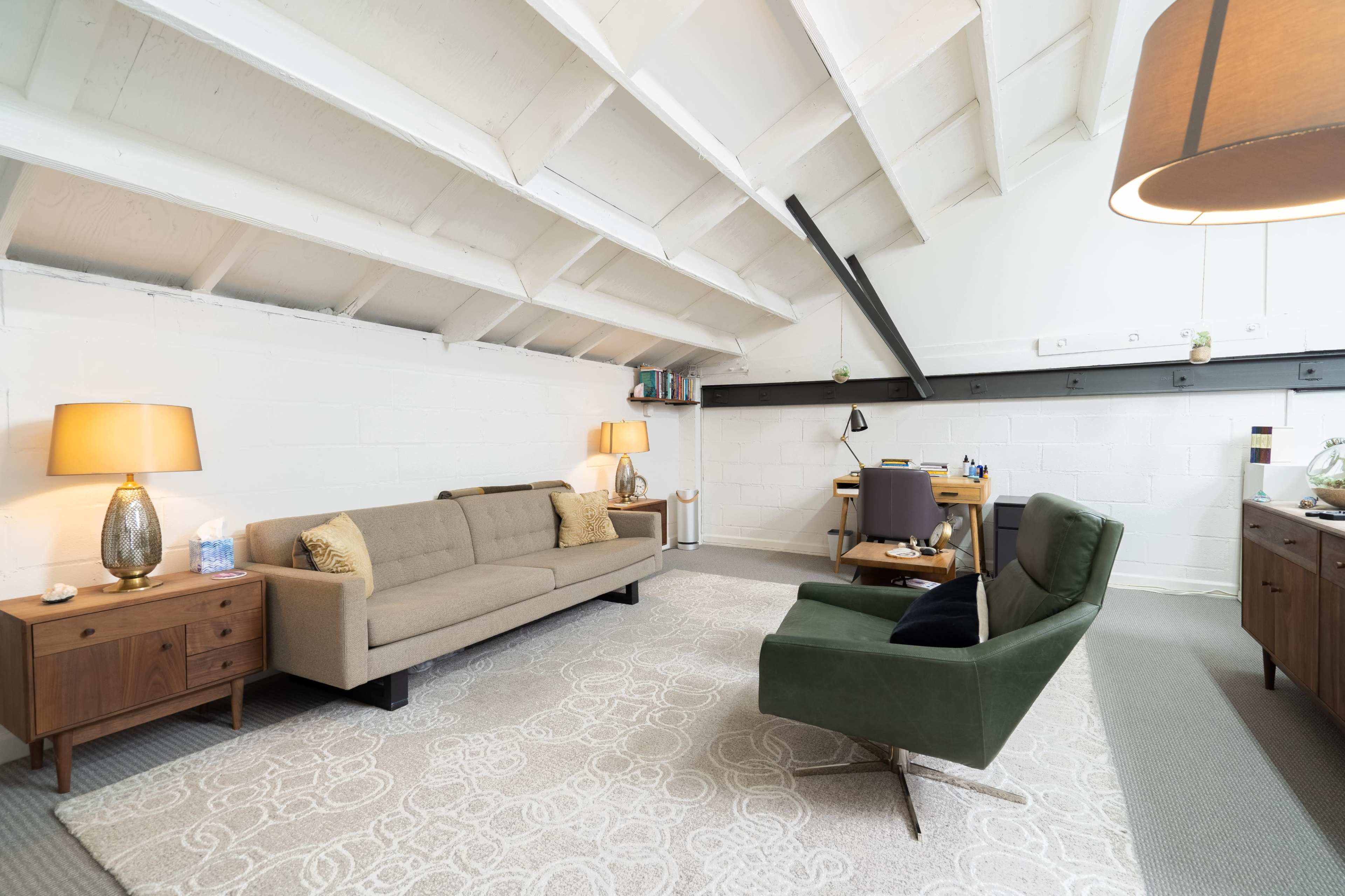 A modern, minimalistic room with a beige sofa, a green armchair, and a desk under a sloped ceiling.