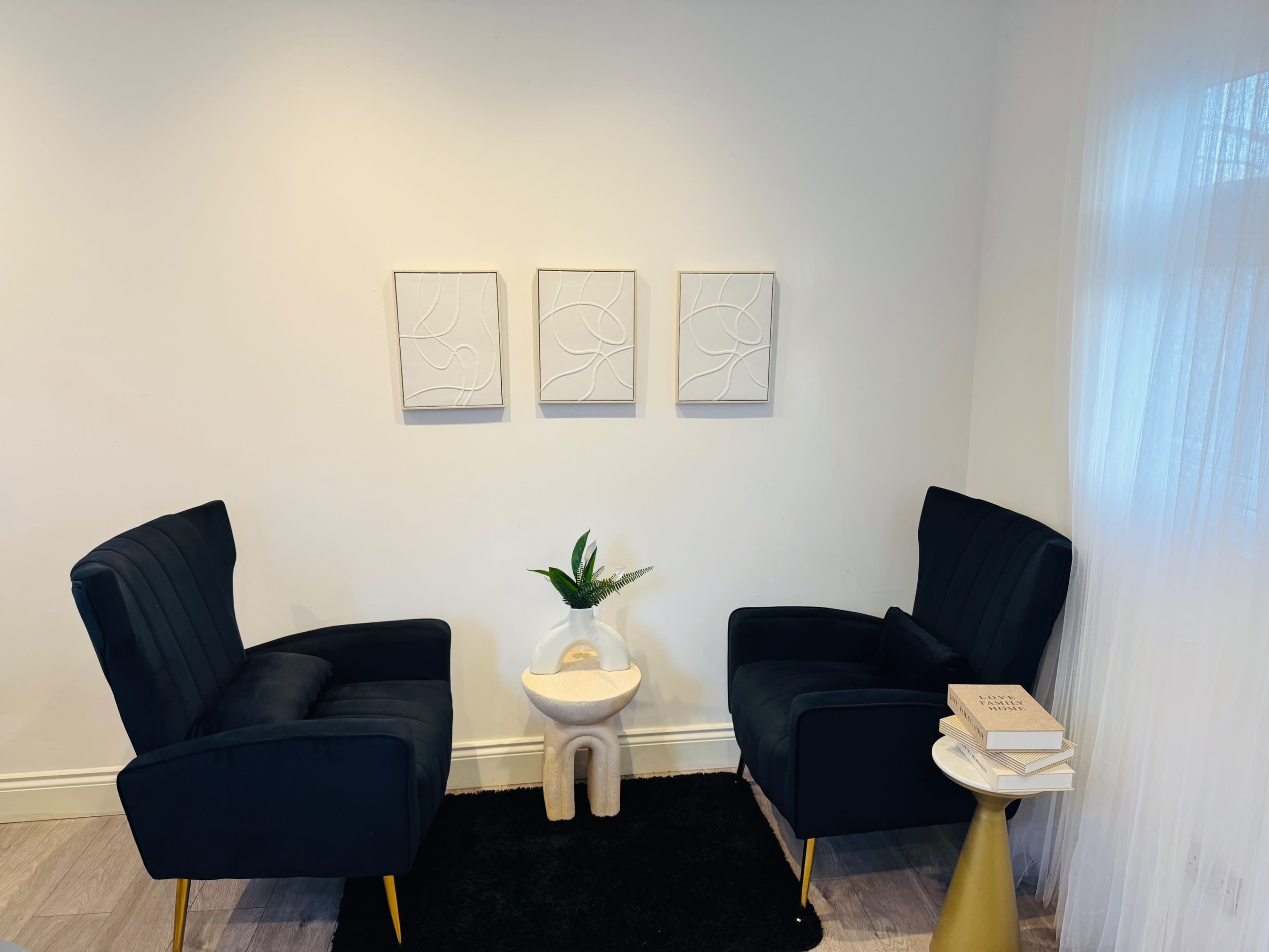 The image shows a small, cozy corner with two black armchairs facing each other, a rounded table with a plant and books in the center, and three framed wall art pieces above.