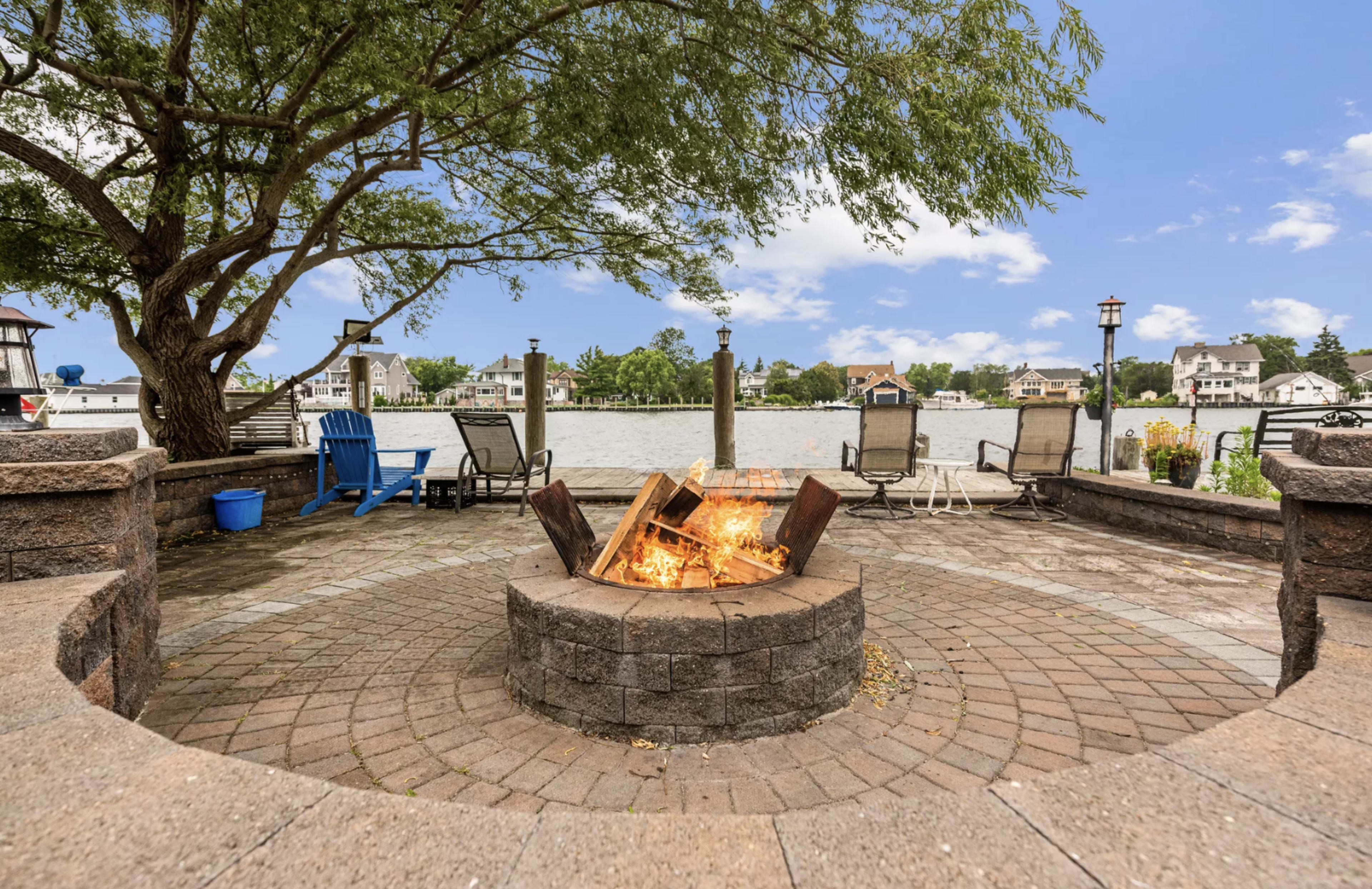A fire pit with flames is surrounded by seating and overlooks a calm lake with houses in the background.