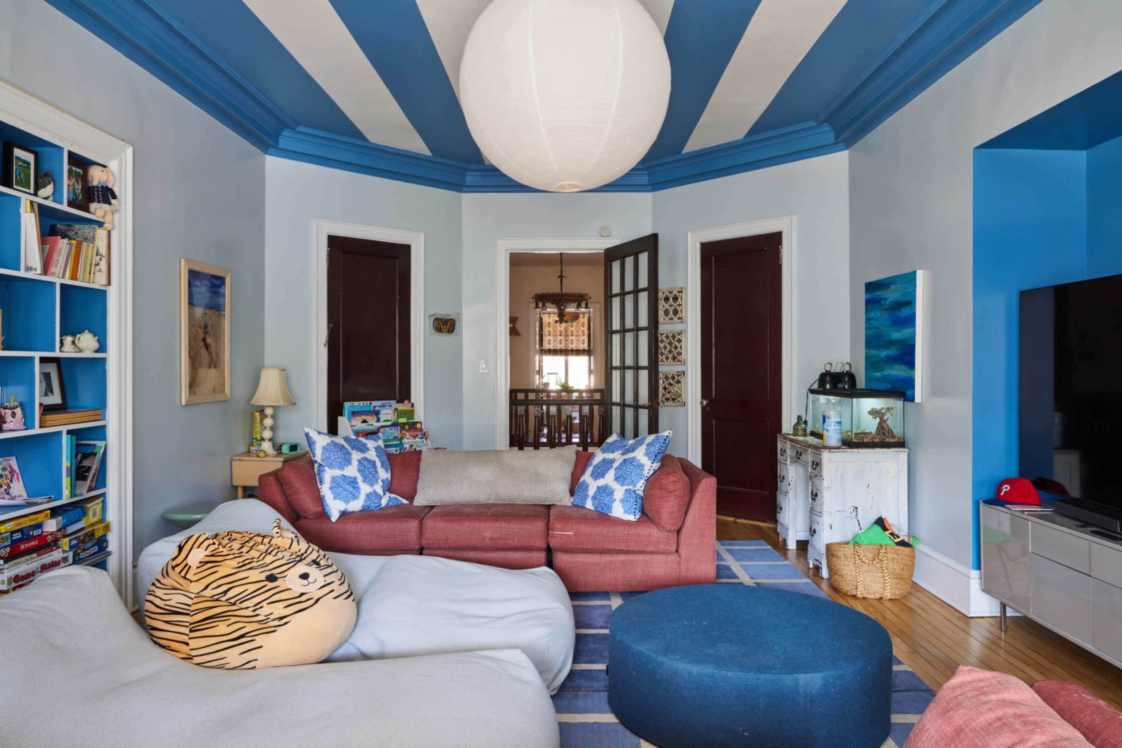 A cozy living room features a sectional sofa, patterned cushions, a large bean bag, and a circular rug, with blue-striped ceilings and a lantern-style light fixture.