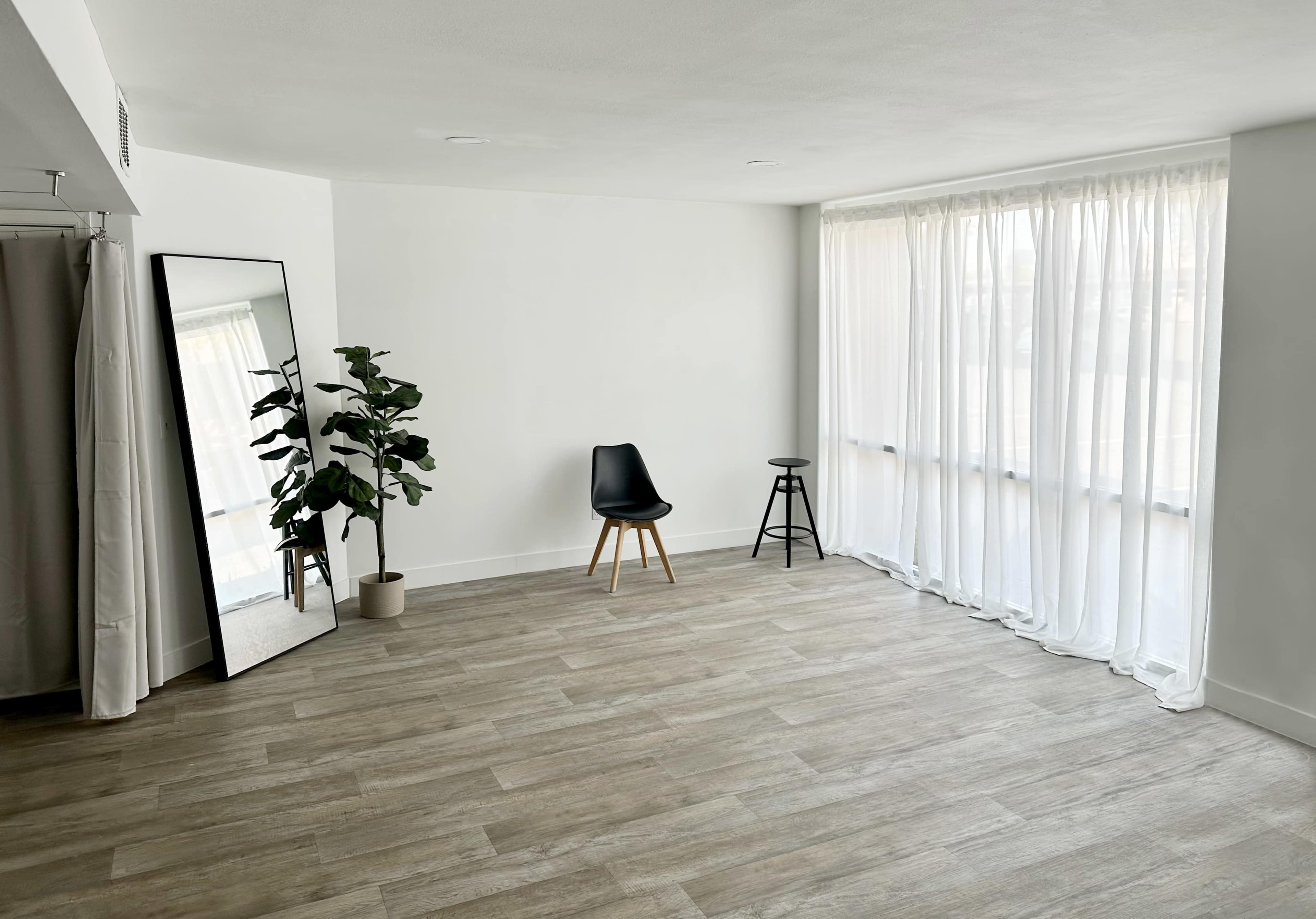 A spacious room features a large mirror, a potted plant, a black chair, and a stool, with bright daylight streaming through sheer curtains.