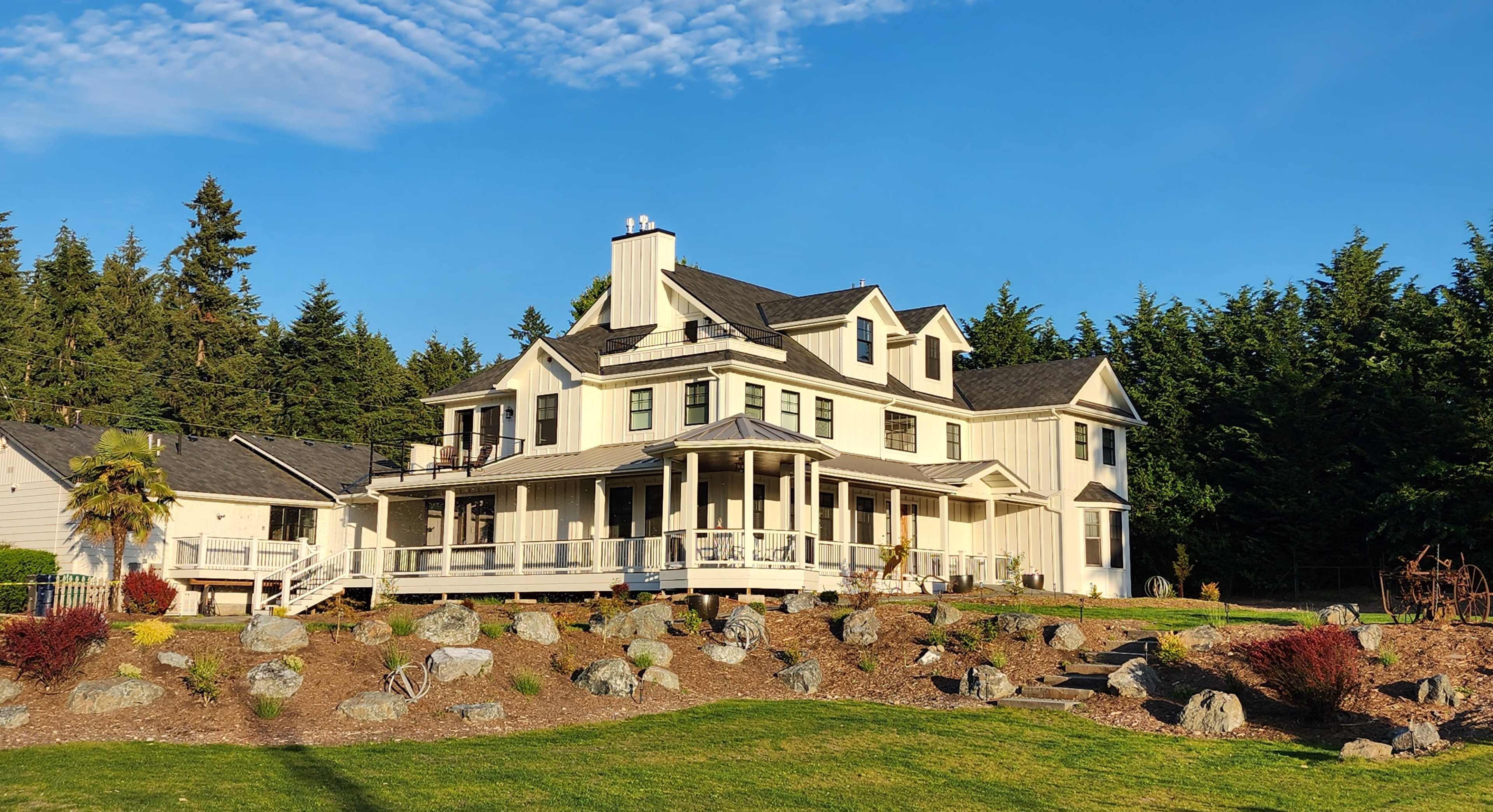 A large white house with multiple gables and a wraparound porch is surrounded by landscaped rocks and greenery on a clear day.